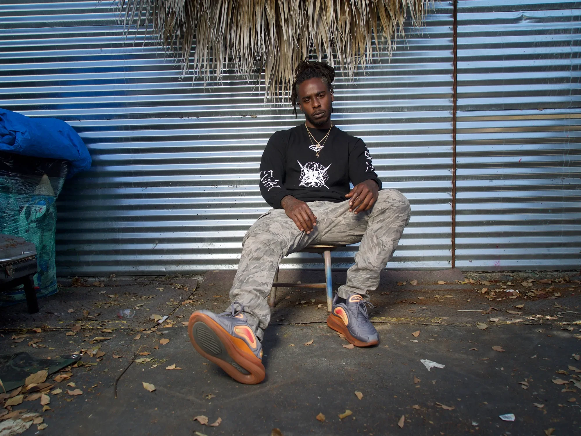 A young man with dreadlocks sits on a stool against a corrugated metal wall. He wears a black graphic long-sleeve shirt, camouflage pants, and sneakers, with jewelry including necklaces. Leaves and debris are scattered on the ground.