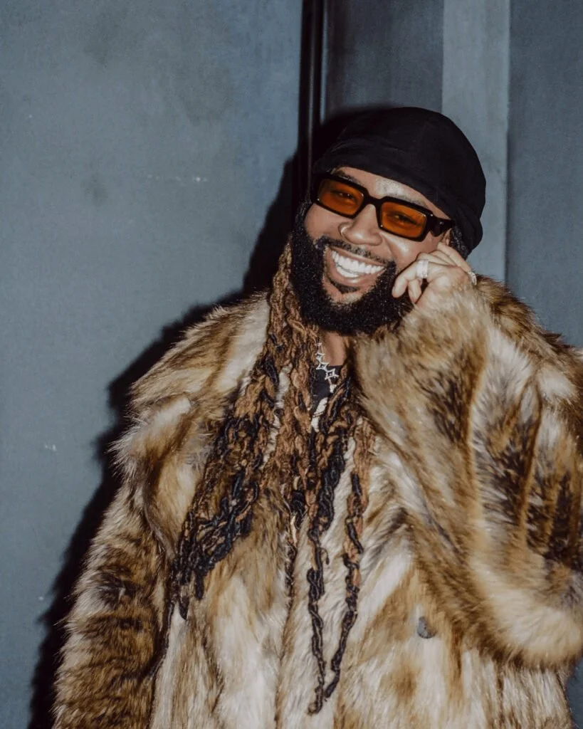A smiling man with a beard and dreadlocks wearing a fur coat, black bandana, and orange-tinted sunglasses, standing against a gray background.