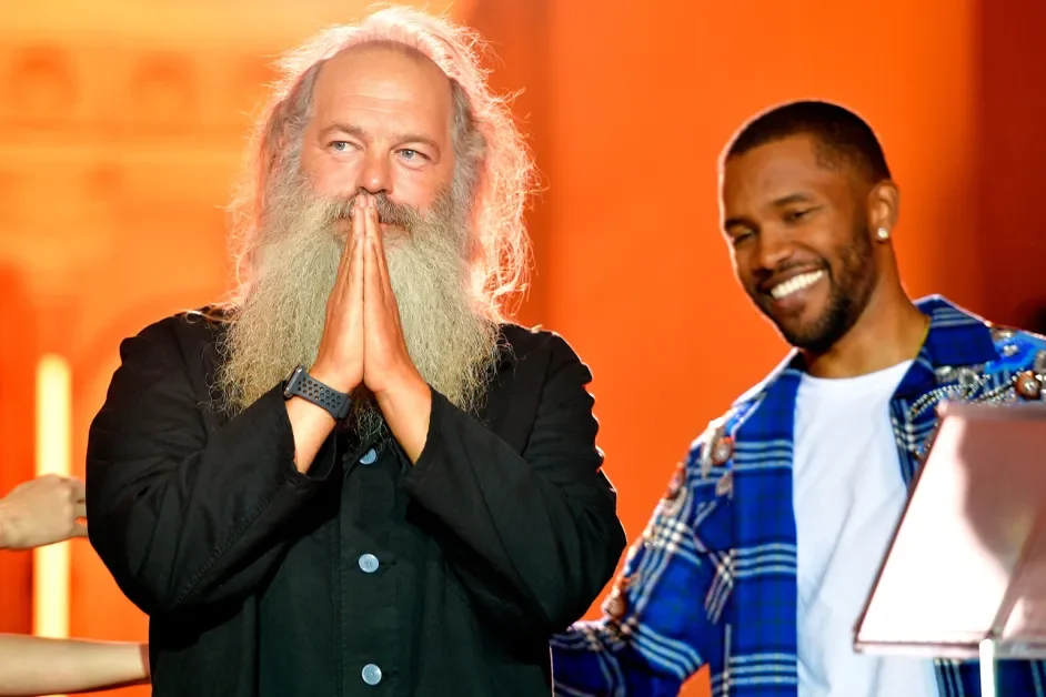 Two men smiling, one with long white hair and beard, the other with short black hair and beard, in an indoor setting with warm orange lighting.