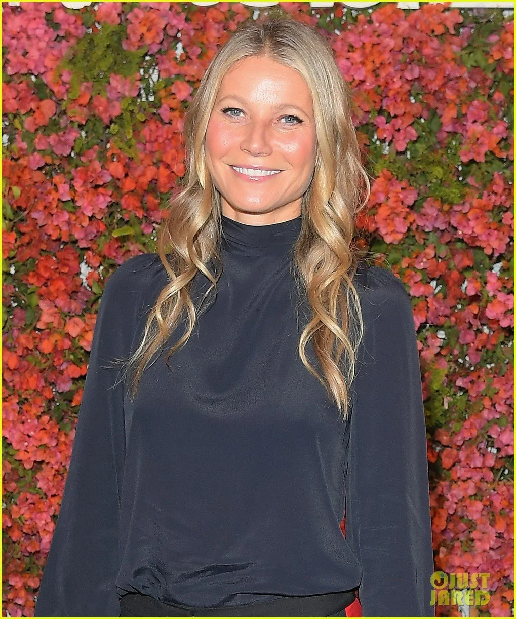 A smiling blonde woman with wavy hair wearing a black turtleneck standing in front of a colorful wall of pink, red, and green flowers.