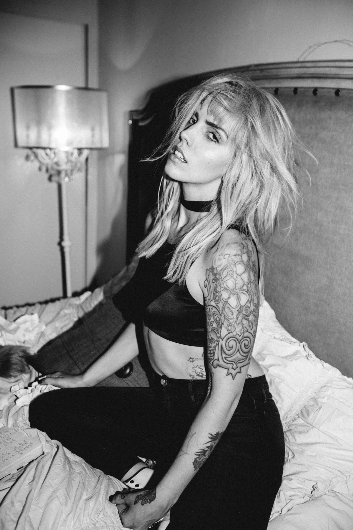 A woman with tattoos on her arm and midriff, blonde hair, and a black choker, sitting on a bed with disheveled sheets, in a room with a lamp and headboard in the background.