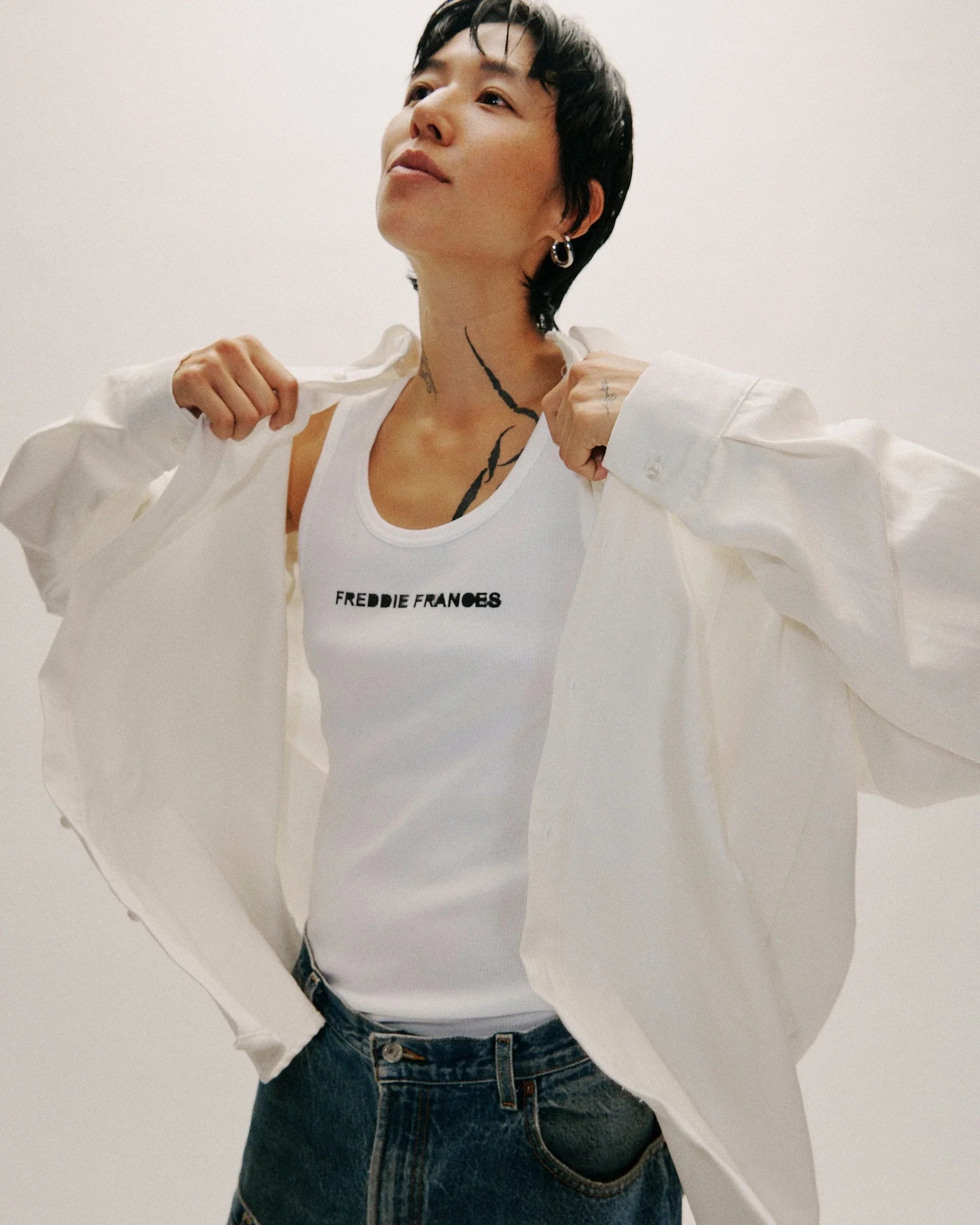 Person wearing a white tank top with the text 'FREDIE FRANIES', a white shirt, and jeans, with short black hair, earrings, and a tattoo on their neck, standing against a plain background.