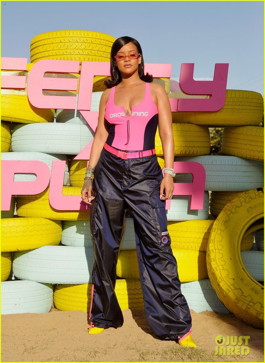 Woman wearing a pink and black jumpsuit and yellow shoes standing in front of colorful stacks of tires in pink, white, and yellow with pink block letters spelling 'EVERY' in the background.
