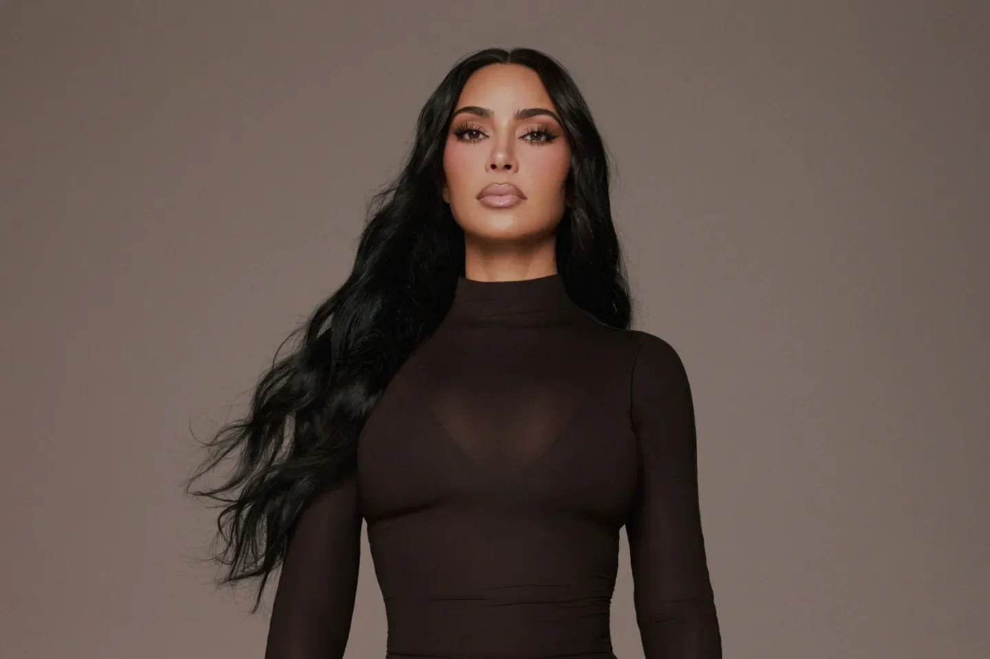 A woman with long wavy black hair, wearing a black long-sleeve top, standing against a neutral gray background.