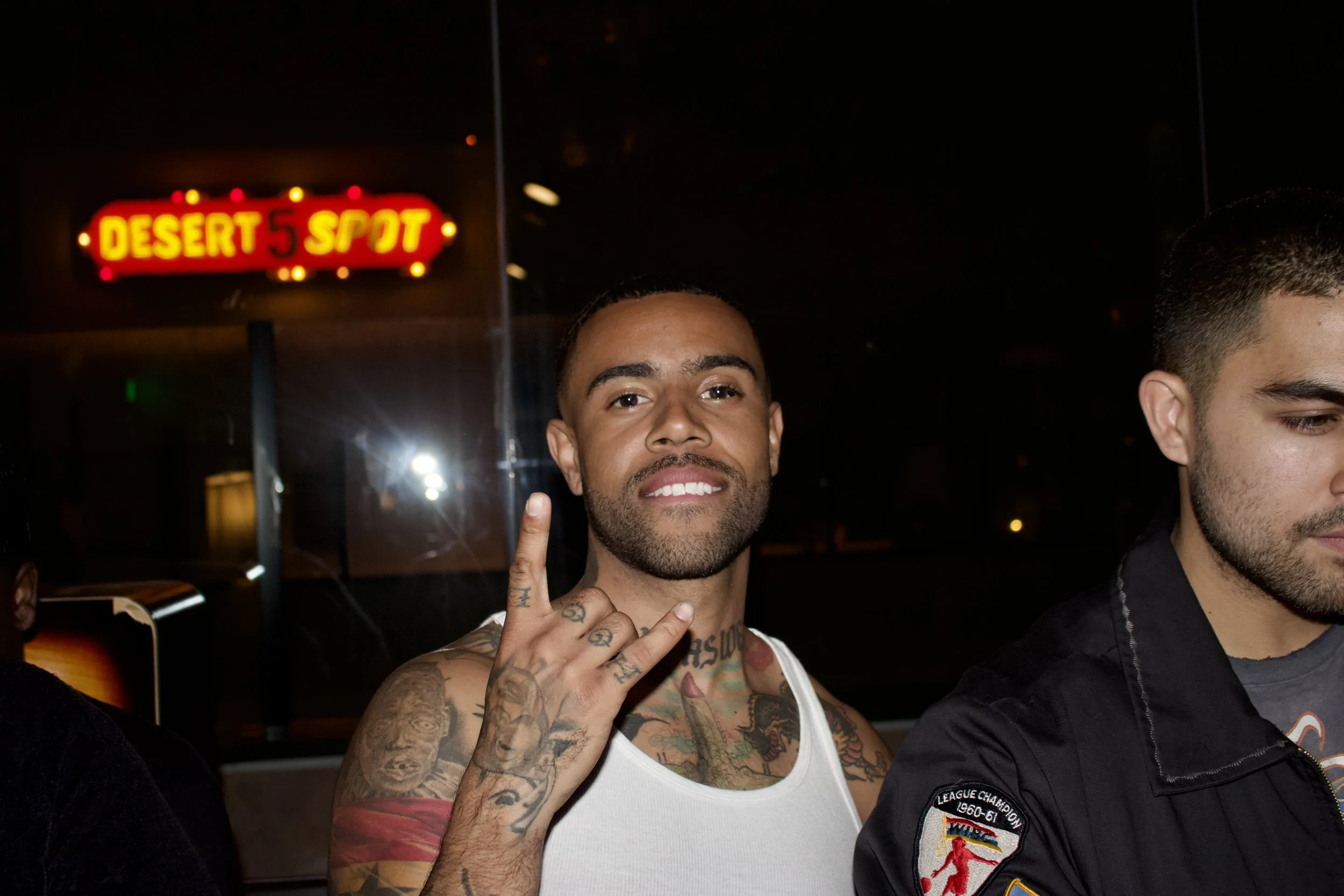A man with tattoos and a white tank top making a hand sign with his fingers at a nightclub or bar, with a red neon sign reading 'Desert 5 Spot' in the background.