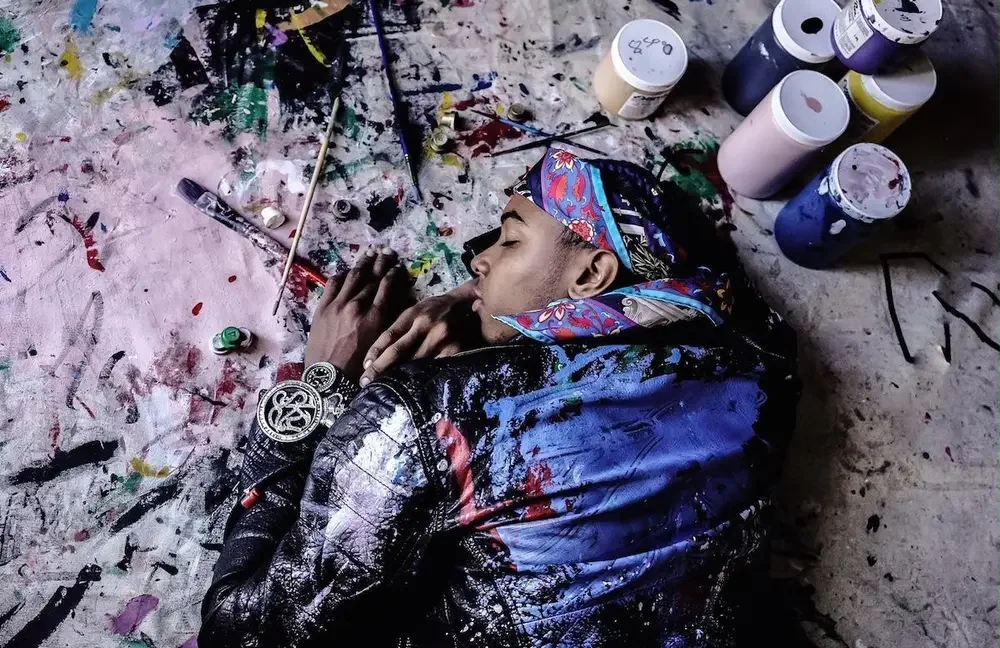 A person lying on the ground in an art studio surrounded by paint bottles and brushes, appearing to be asleep or resting.
