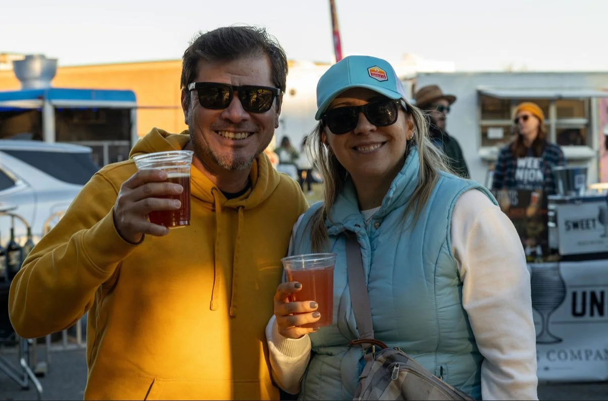 Two people smiling and holding cups of iced drinks at an outdoor event, wearing sunglasses and casual clothing.
