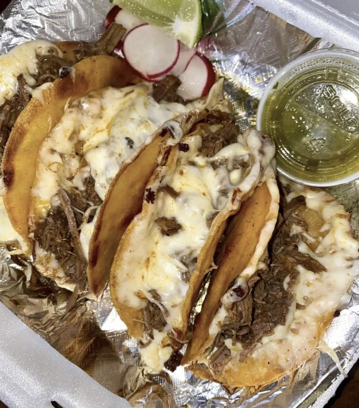 Three authentic Mexican tacos with shredded beef, cheese, and queso fresco, topped with radishes, lime wedge, and a small cup of green salsa, served on foil-lined plate.