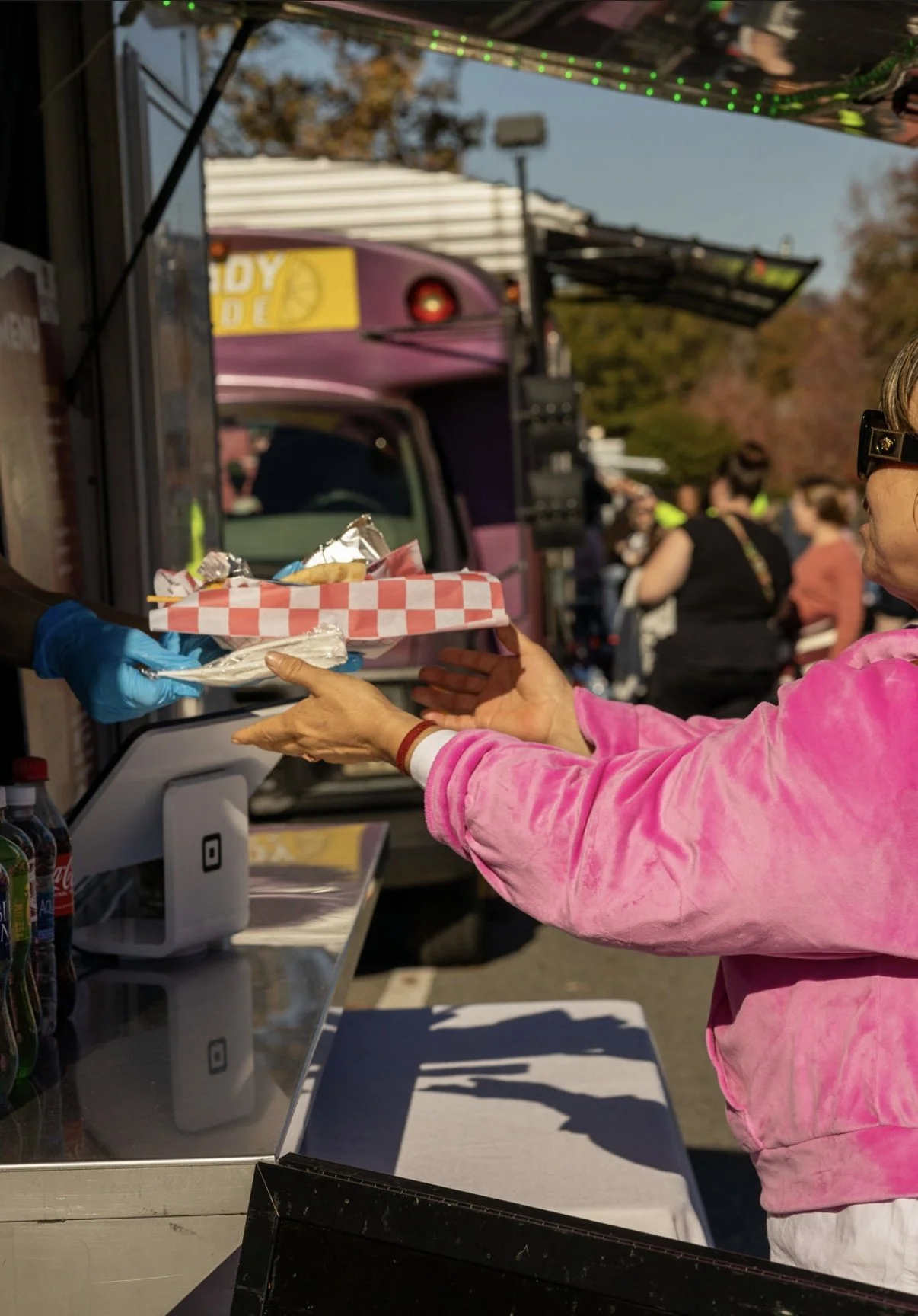 A woman in a pink jacket receiving food in a checkered tray from a person wearing blue gloves at an outdoor food stand, with a colorful food truck and a crowd in the background.