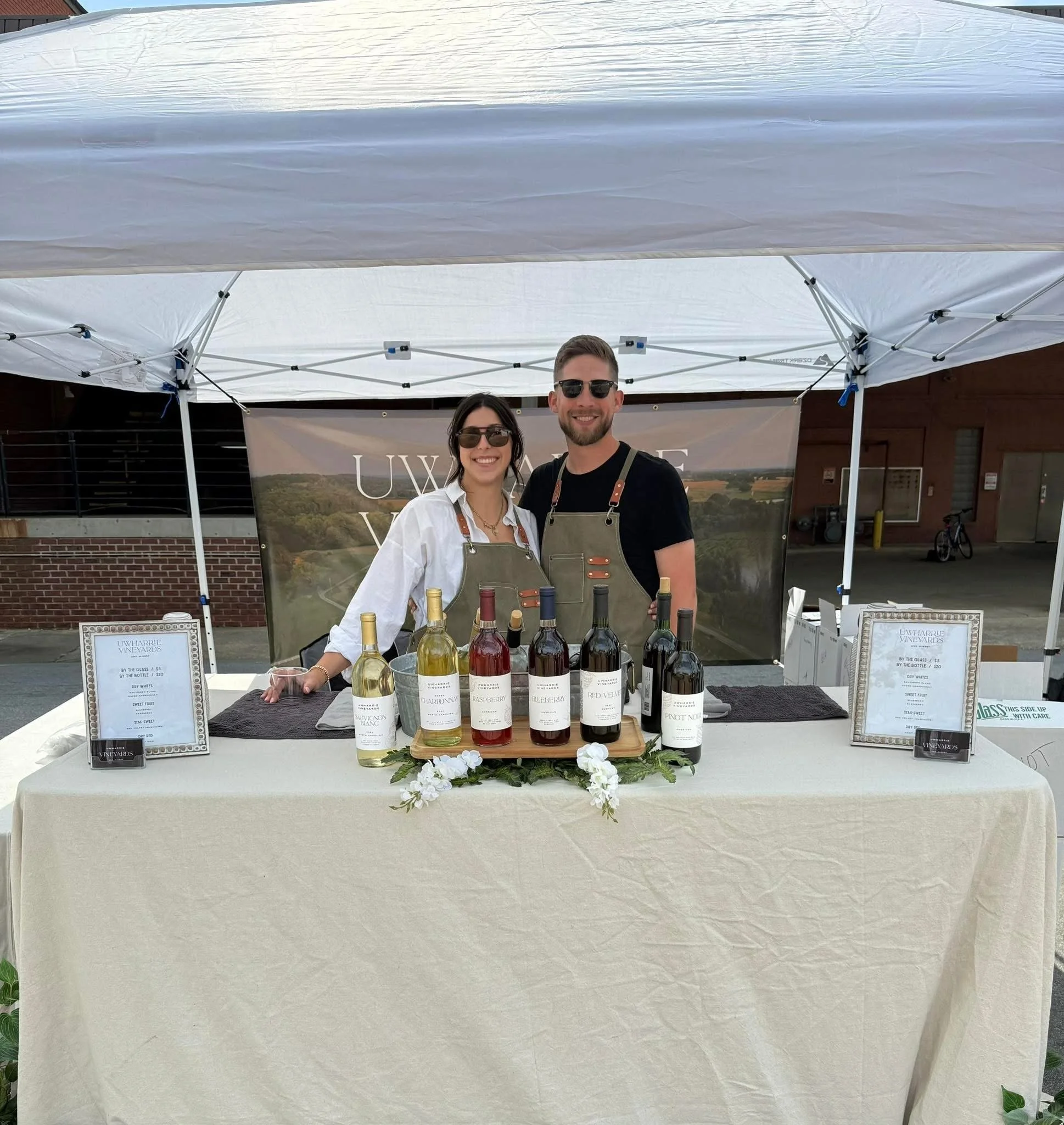 A man and woman behind a table with wine bottles at an outdoor vineyard event, both wearing aprons and sunglasses, smiling at the camera.