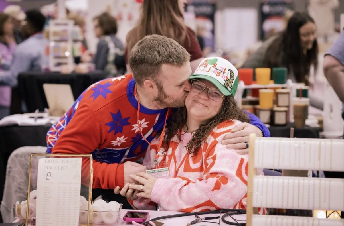 A man with short hair and a beard kissing a woman with glasses and long curly hair on the cheek at a busy indoor event. The woman is smiling with her eyes closed, wearing a colorful hat and a patterned sweater. The background features tables with various items and other people.