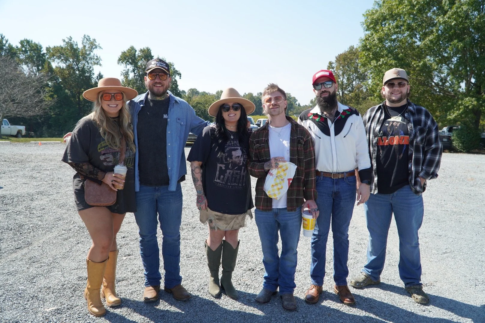 Group of six people standing outdoors on a sunny day, smiling, holding drinks, with trees and parked cars in the background.