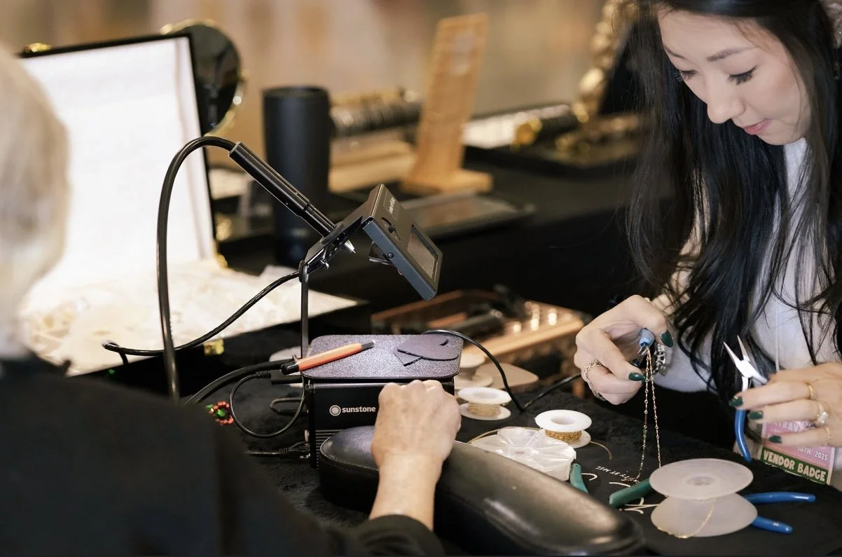 A woman working on jewelry making at a work table with tools, spools of wire, and a magnifier light, in a craft or jewelry studio.