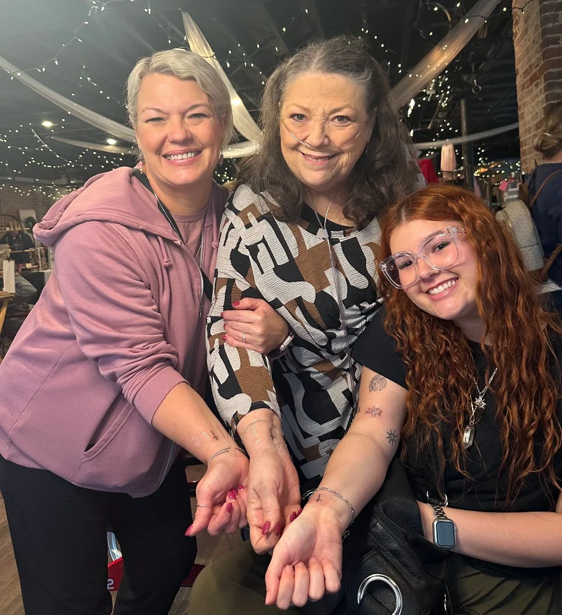 Three women smiling and holding hands at a gathering or celebration, with fairy lights and decorations in the background.