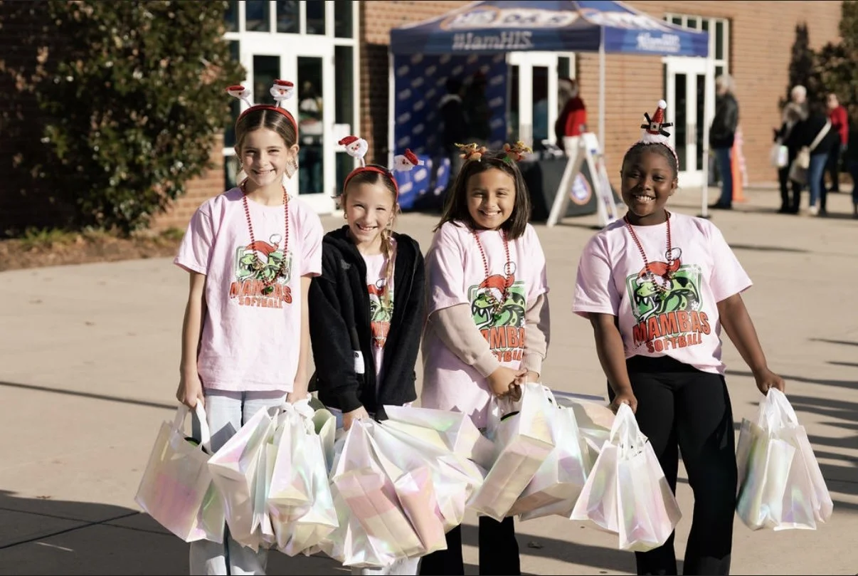 Four young girls smiling and wearing Christmas-themed headbands and neighborhood sports event t-shirts, holding gift bags, standing outdoors near a brick building during daytime.