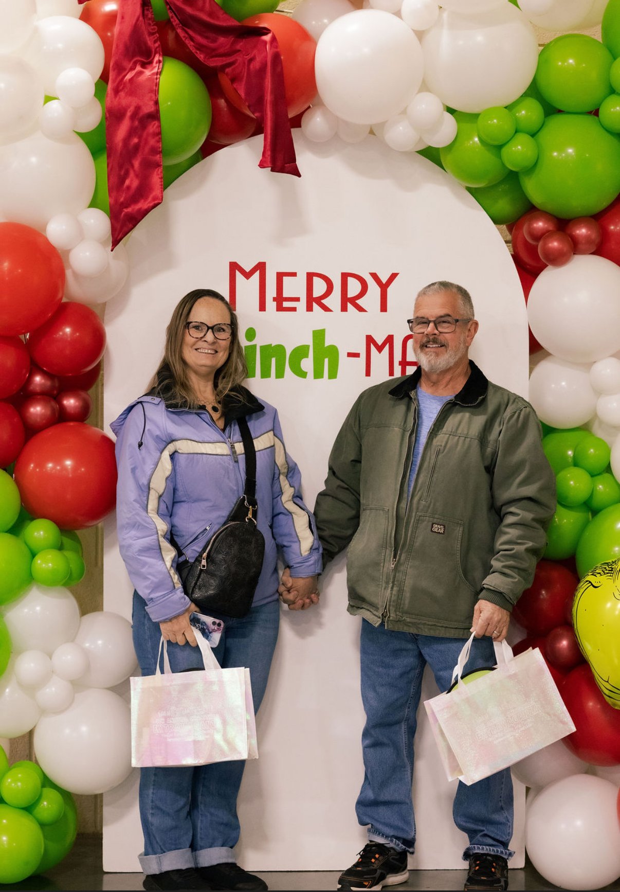 A smiling couple holding hands in front of a festive holiday balloon display with a sign that says "Merry Inch-Mas," surrounded by green, red, and white balloons.