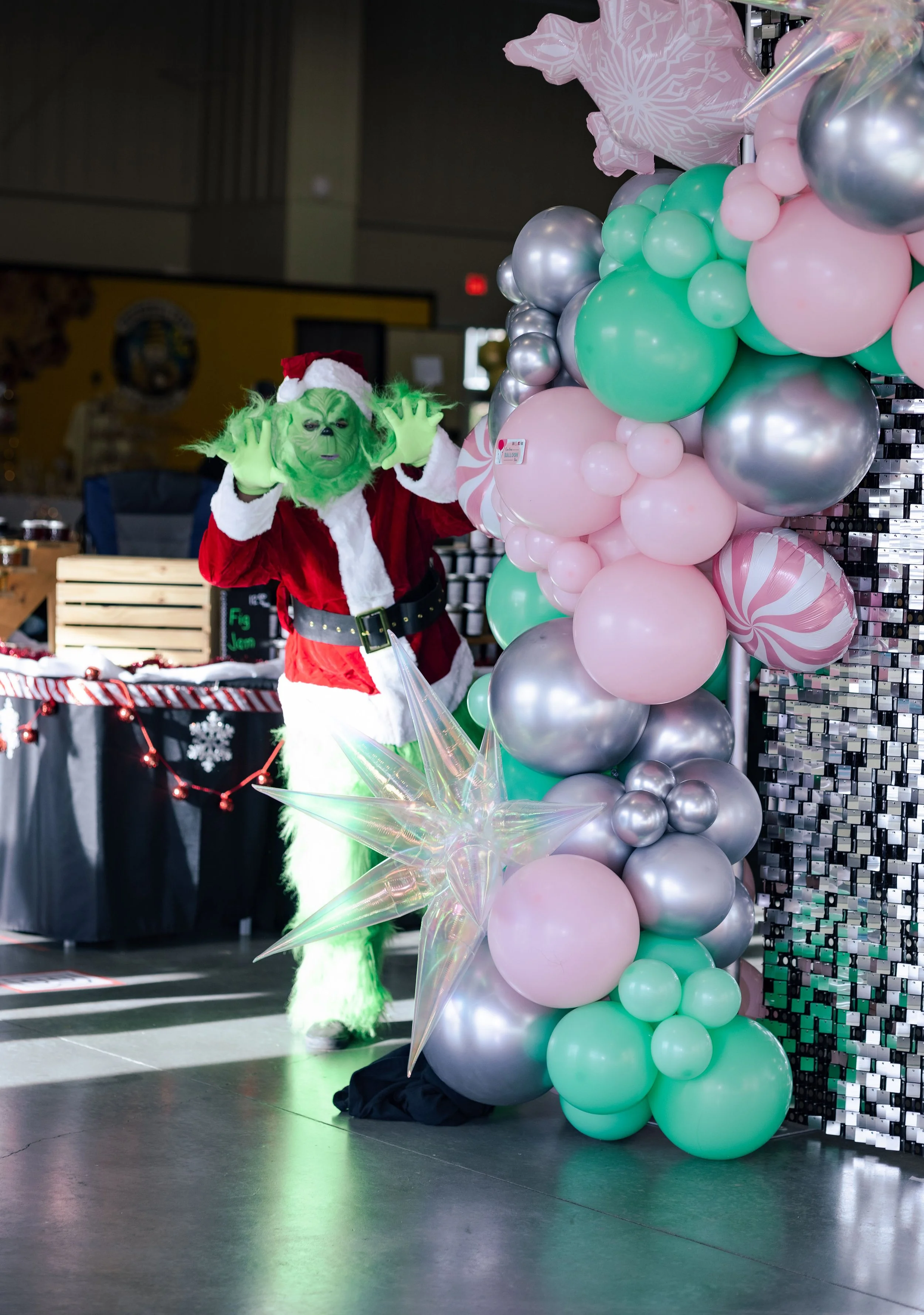 Person dressed as the Grinch wearing a Santa suit, posing next to a large balloon display with pastel pink, green, silver, and patterned balloons at a holiday event.