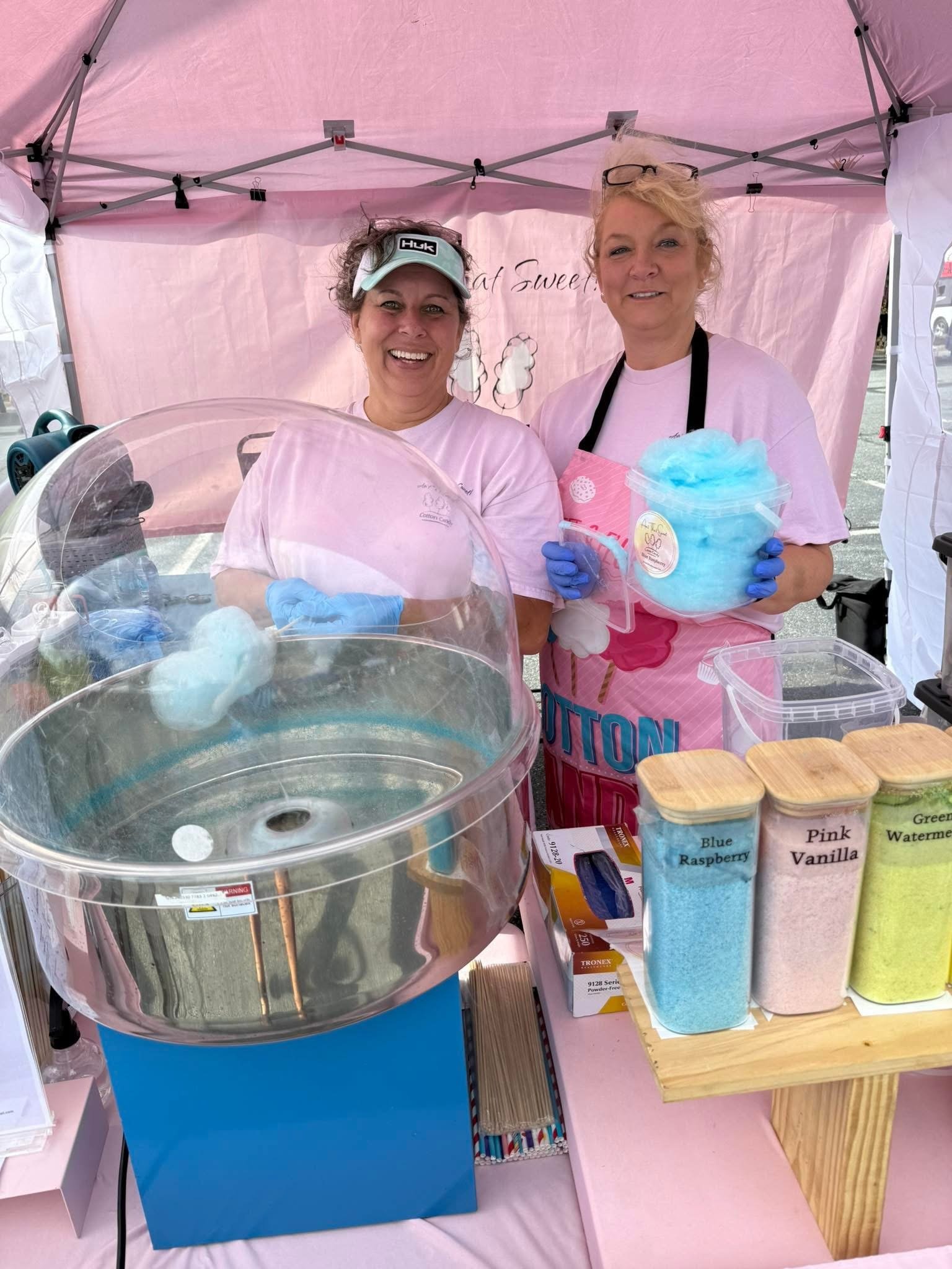 Two women standing behind a pink cotton candy stand, smiling. One woman is holding a container of blue cotton candy, and the other has cotton candy on a stick and is near a cotton candy machine.