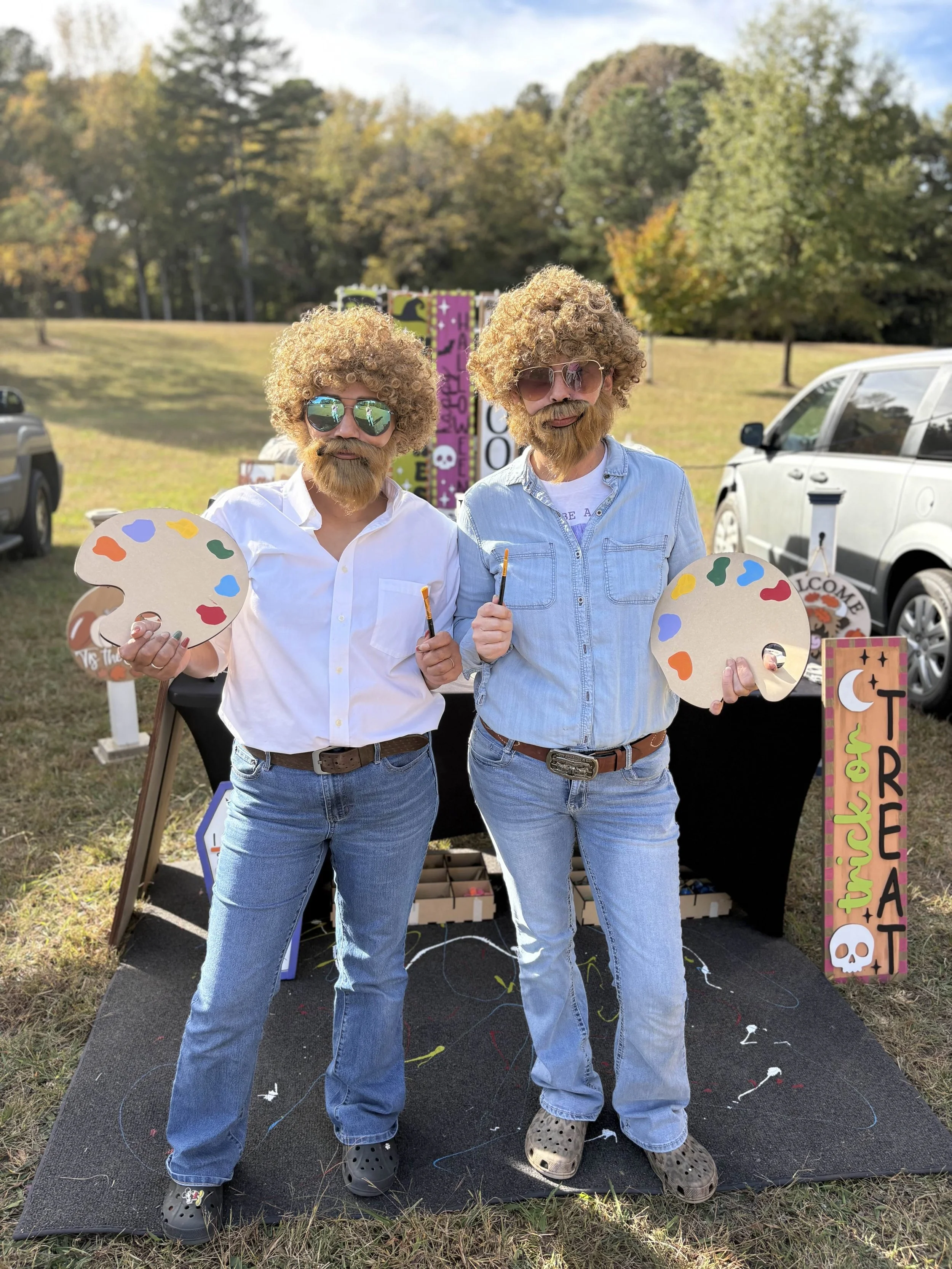 Two men with curly blond hair and beards, wearing sunglasses and casual clothes, holding painter's palettes and brushes, standing outdoors on a grassy field during fall. They are in front of a colorful Halloween-themed display with signs and decorations.