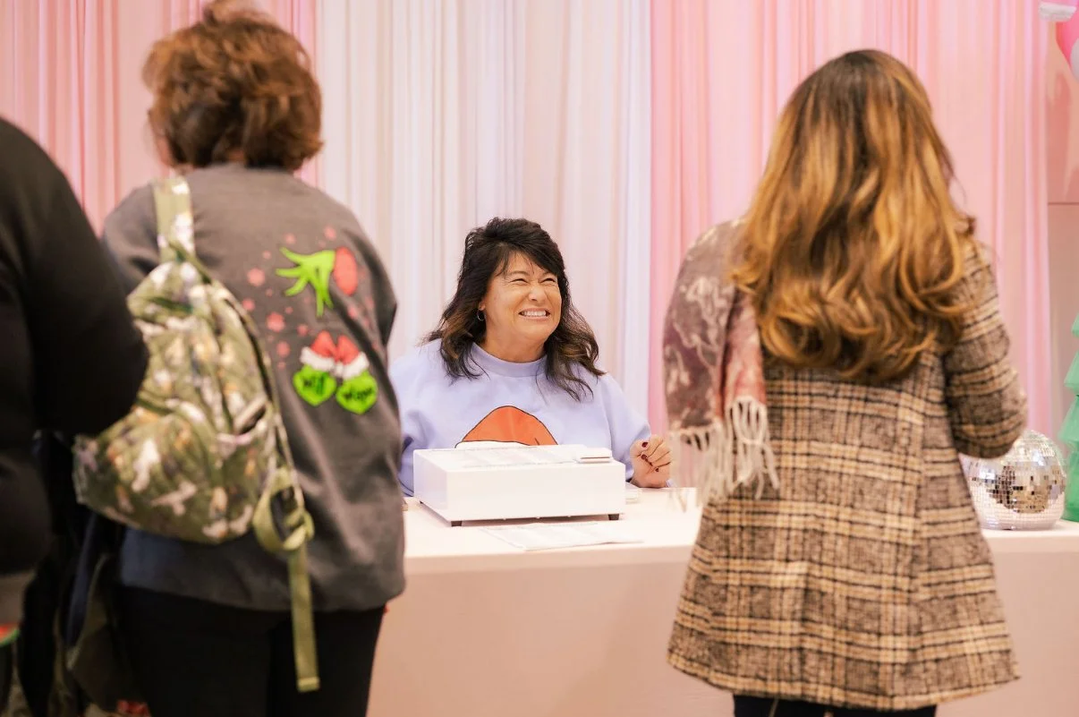 A woman smiling at a table with a cash register, surrounded by three women in festive clothing, in a decorated pink background.