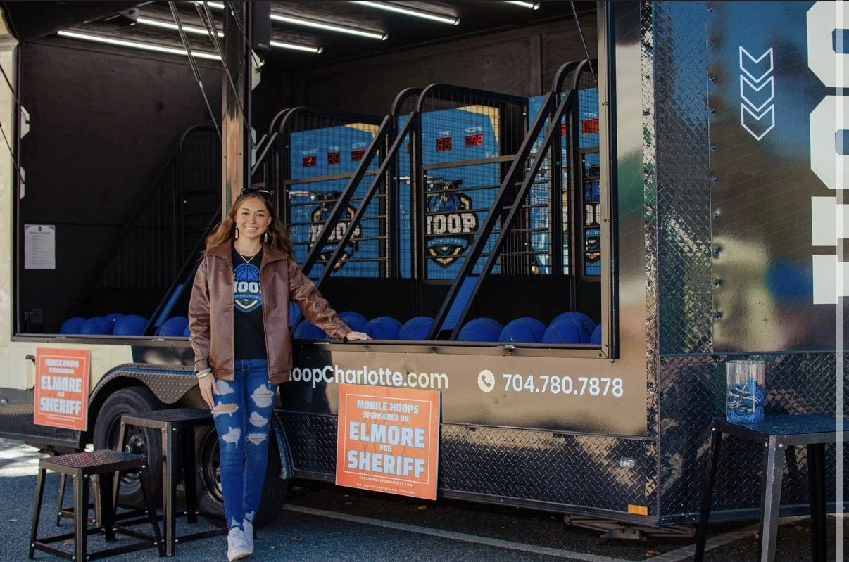 A young woman with long brown hair, wearing a brown leather jacket, a black shirt, and ripped jeans, stands next to a mobile hoop shooting game at an outdoor event sponsored by Elmore County Sheriff. The game features blue hoops and an electronic scoreboard, with a sign displaying a phone number and website. There are two small tables with chairs around the mobile game setup.