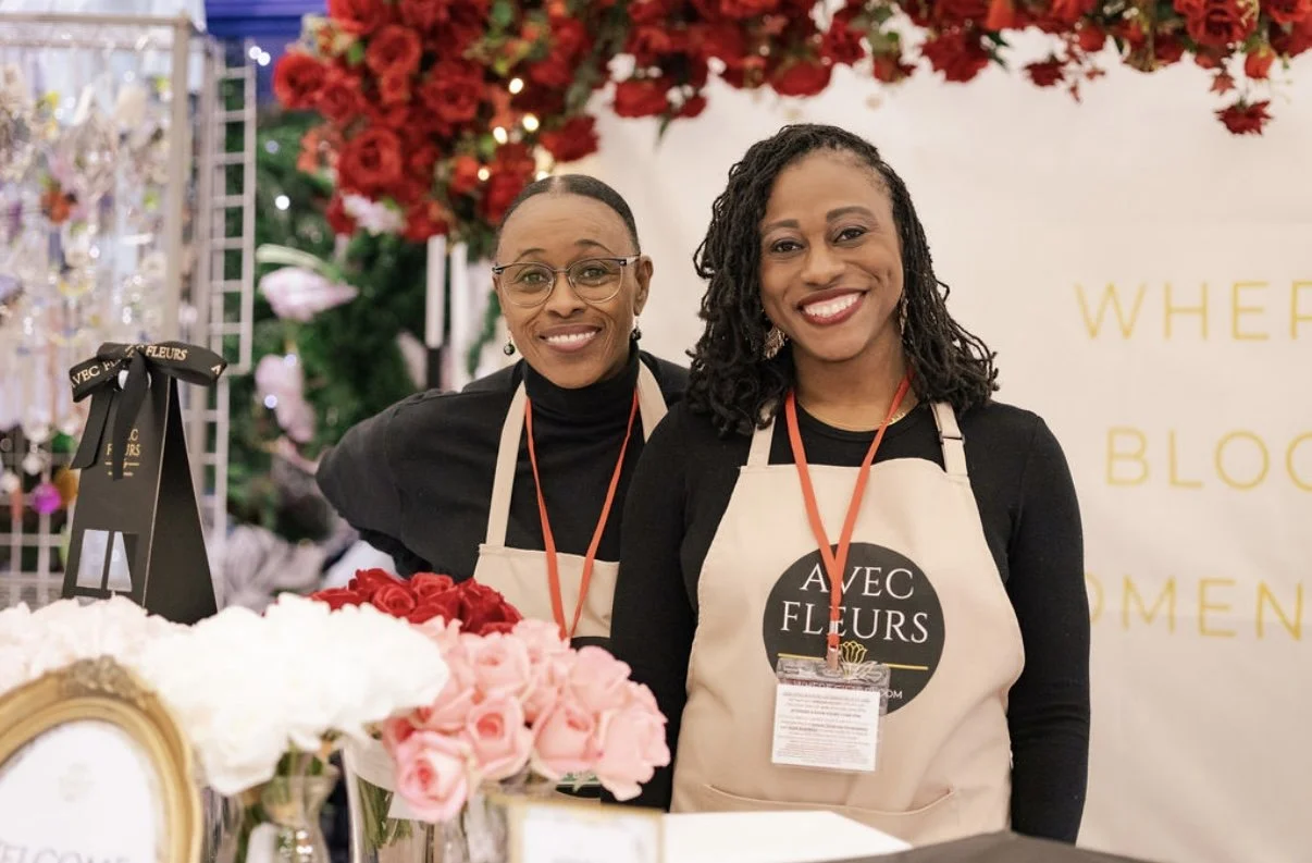 Two women smiling at a floral booth with pink and white roses, red flowers in the background, and a sign that reads 'AVEC FLEURS'.