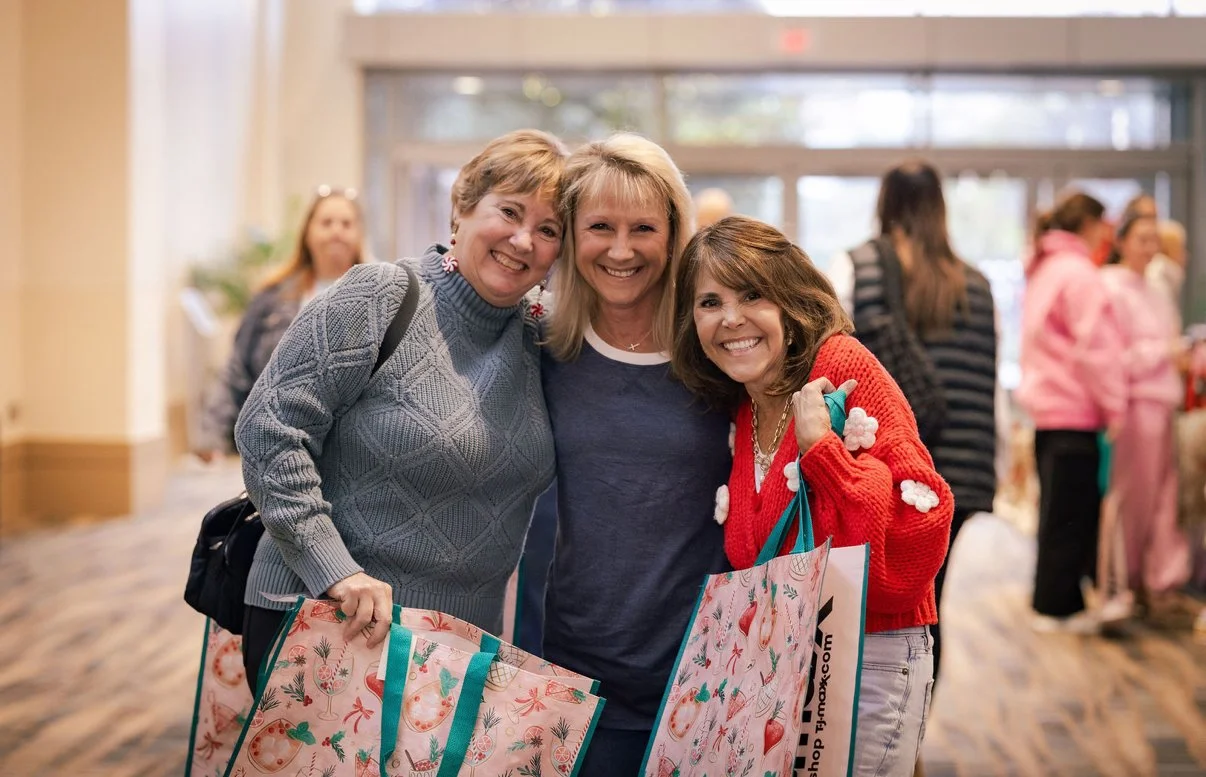 Three women smiling at a gathering, holding shopping bags, in a lively indoor setting with other people in the background.