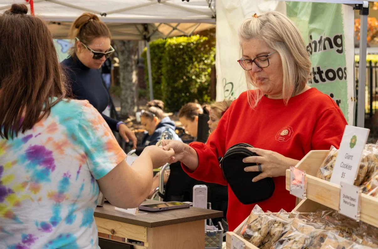 A woman in a red sweatshirt is purchasing cookies from a vendor at an outdoor market, exchanging money.
