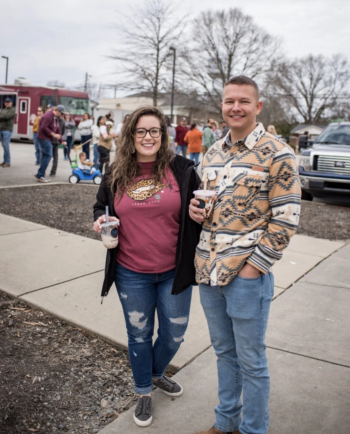 Two smiling people standing outside, holding bubble teas, at what appears to be a community event with other people in the background.