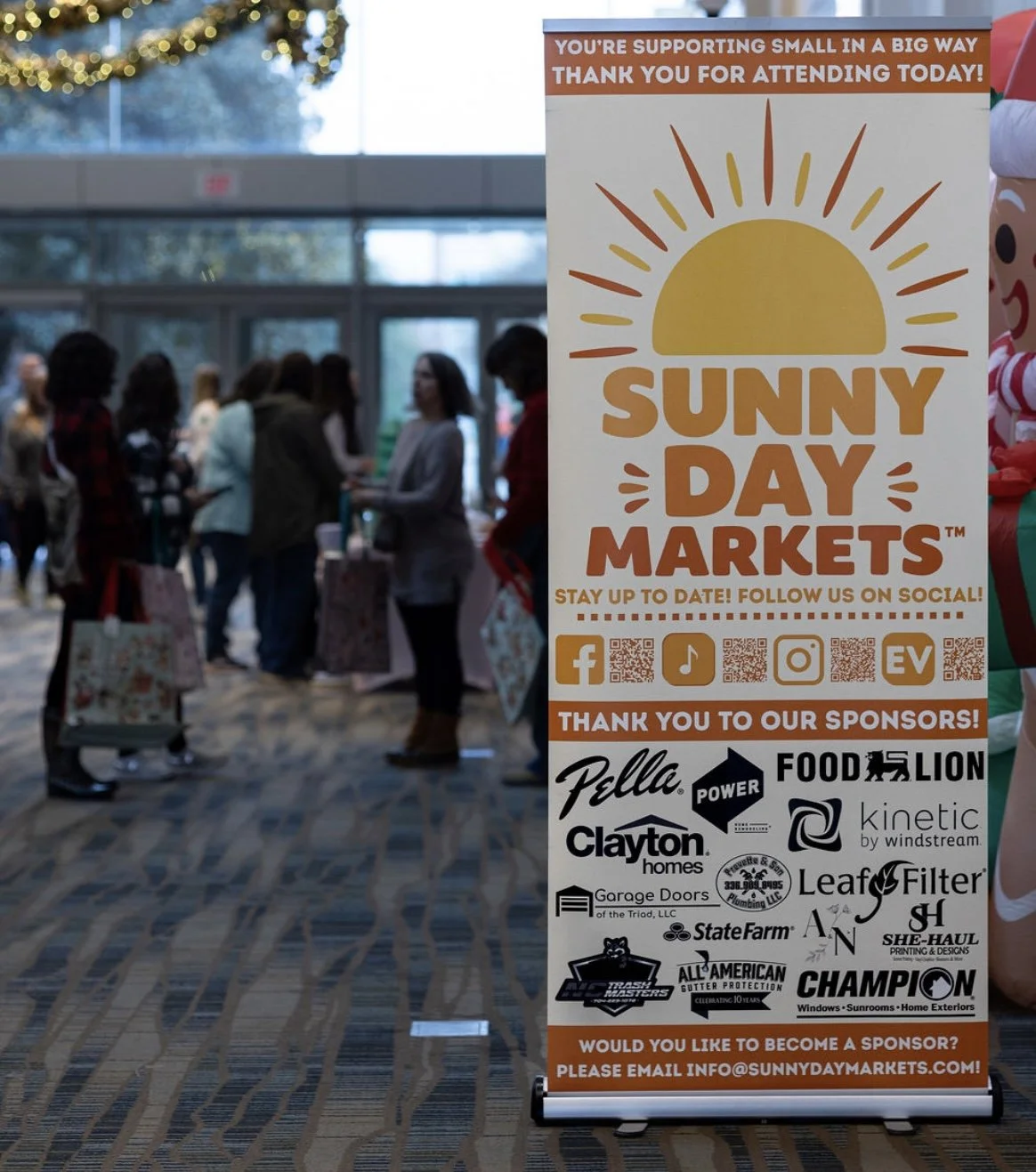 A banner at Sunny Day Markets, supporting small businesses, with logos of various sponsors and social media icons, and a group of people inside a convention center in the background.