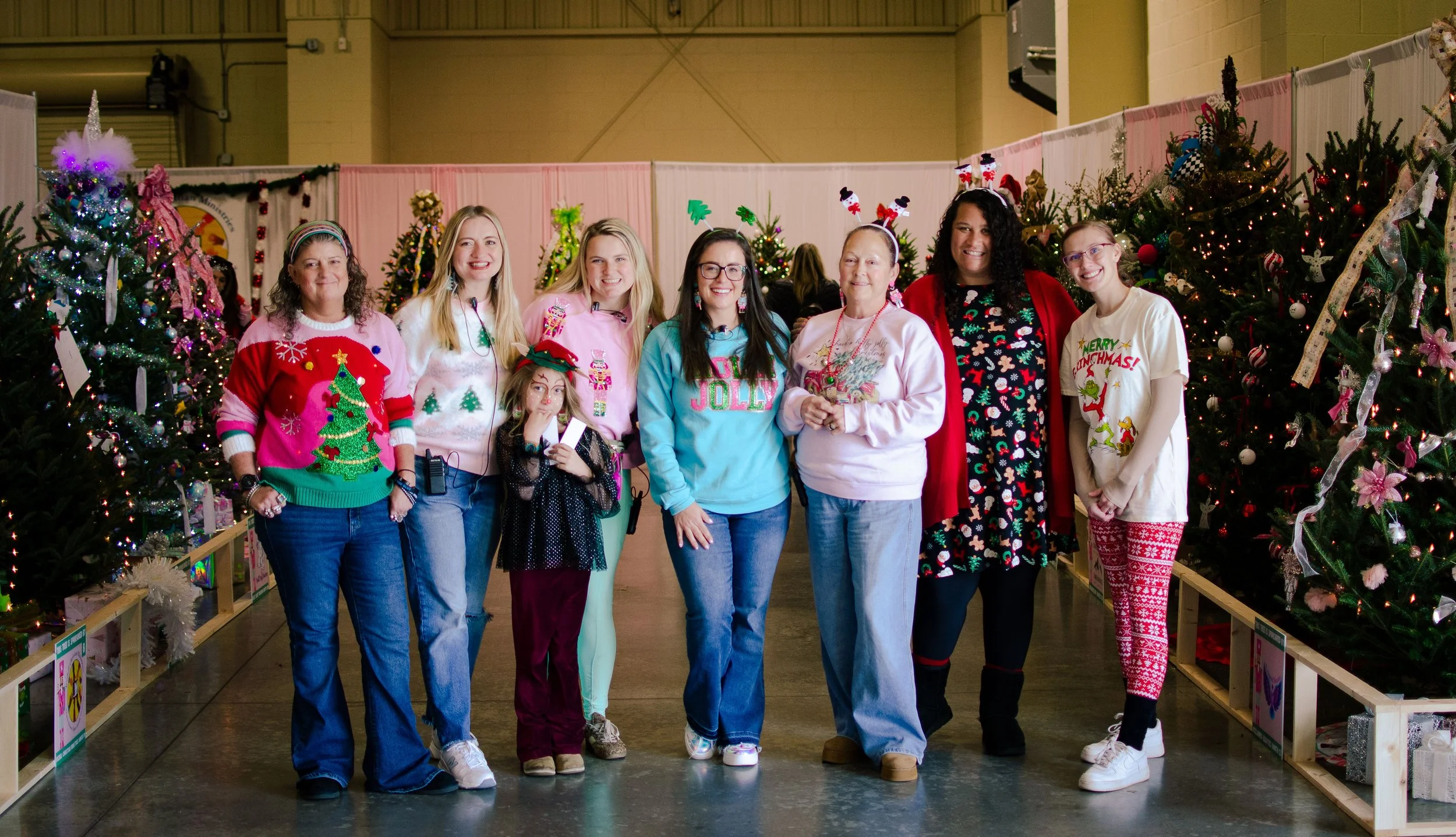 Group of women and girls standing in front of decorated Christmas trees wearing festive holiday sweaters and accessories at a Christmas event.