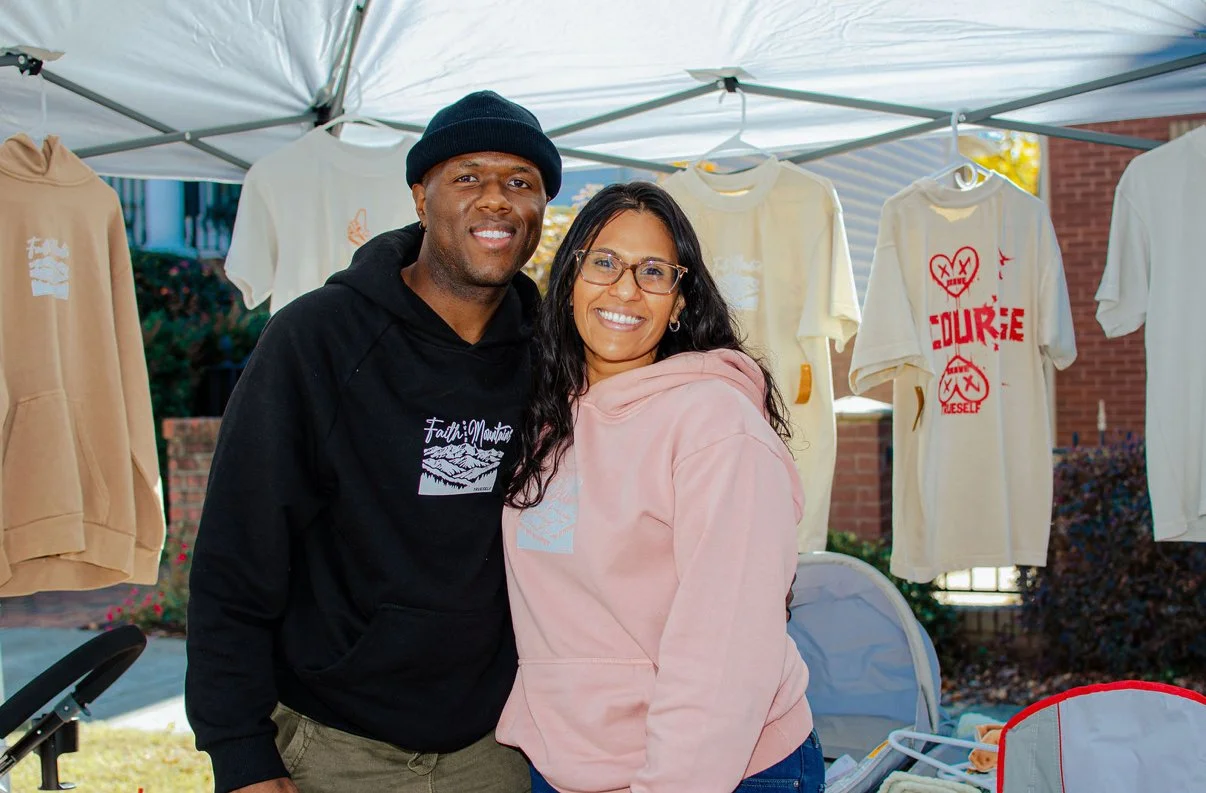 Two smiling people stand close together at a street vendor booth with hanging t-shirts, one man wearing a black hoodie and beanie, and one woman wearing a pink hoodie with glasses.
