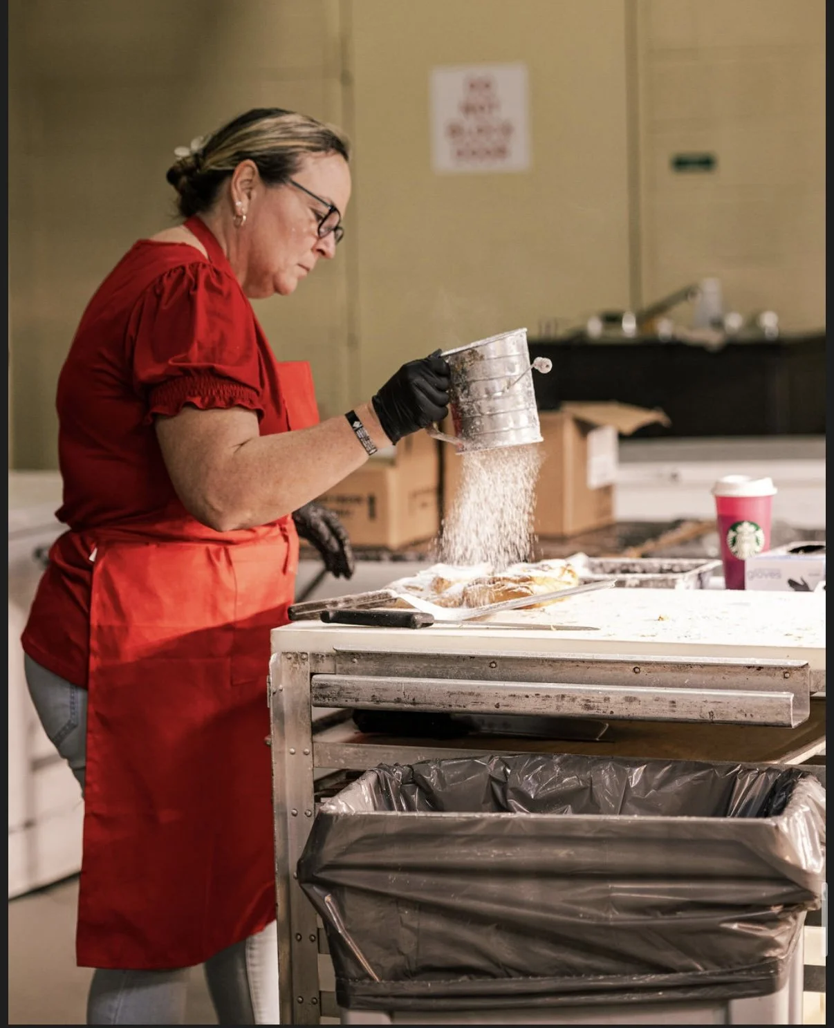 A woman in a red shirt and apron sifting powder onto food on a white table in a kitchen.