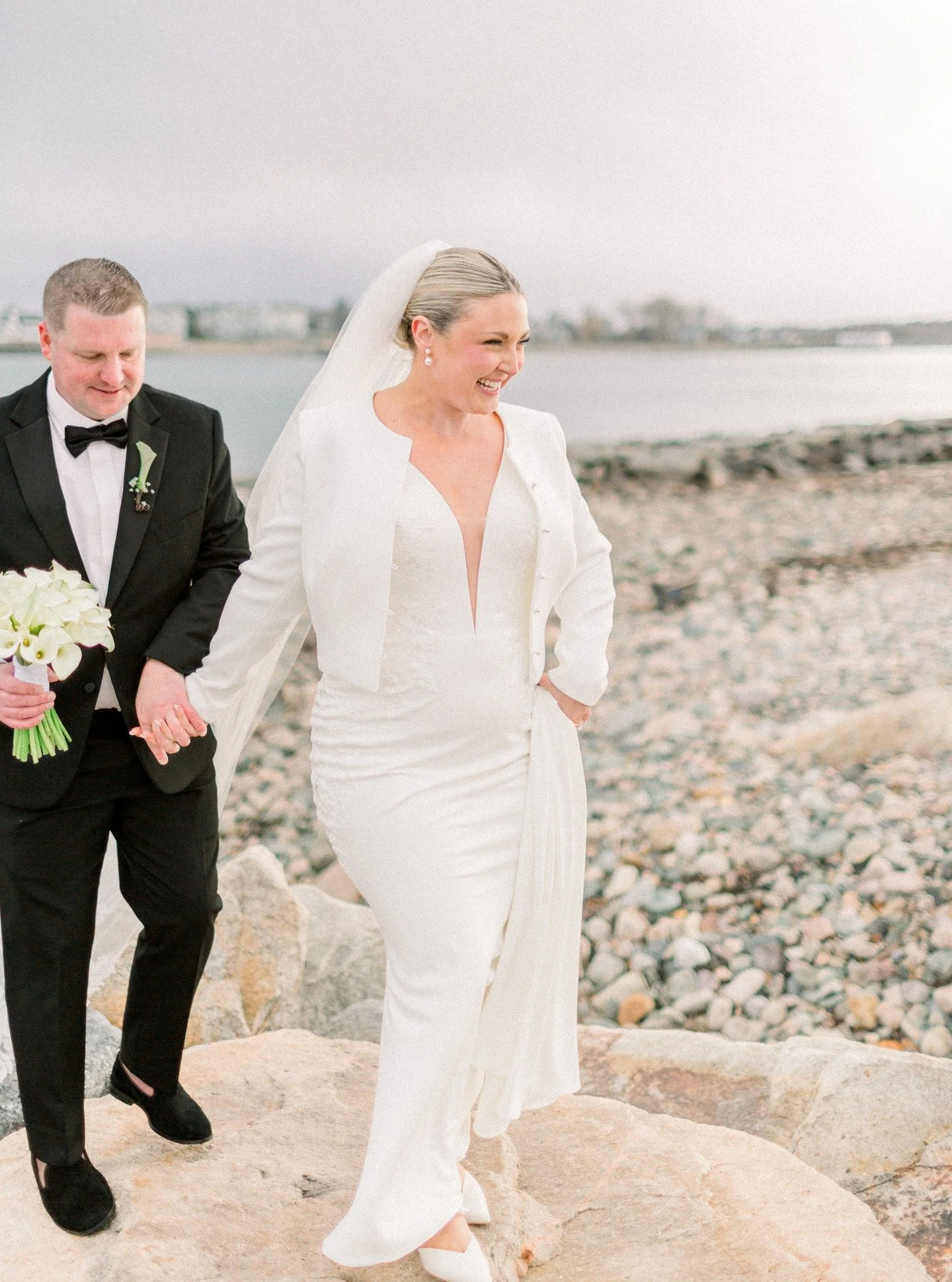 A bride and groom walking on a rocky beach holding hands, with the bride smiling and the groom holding a bouquet of white calla lilies, near a body of water with cloudy skies.