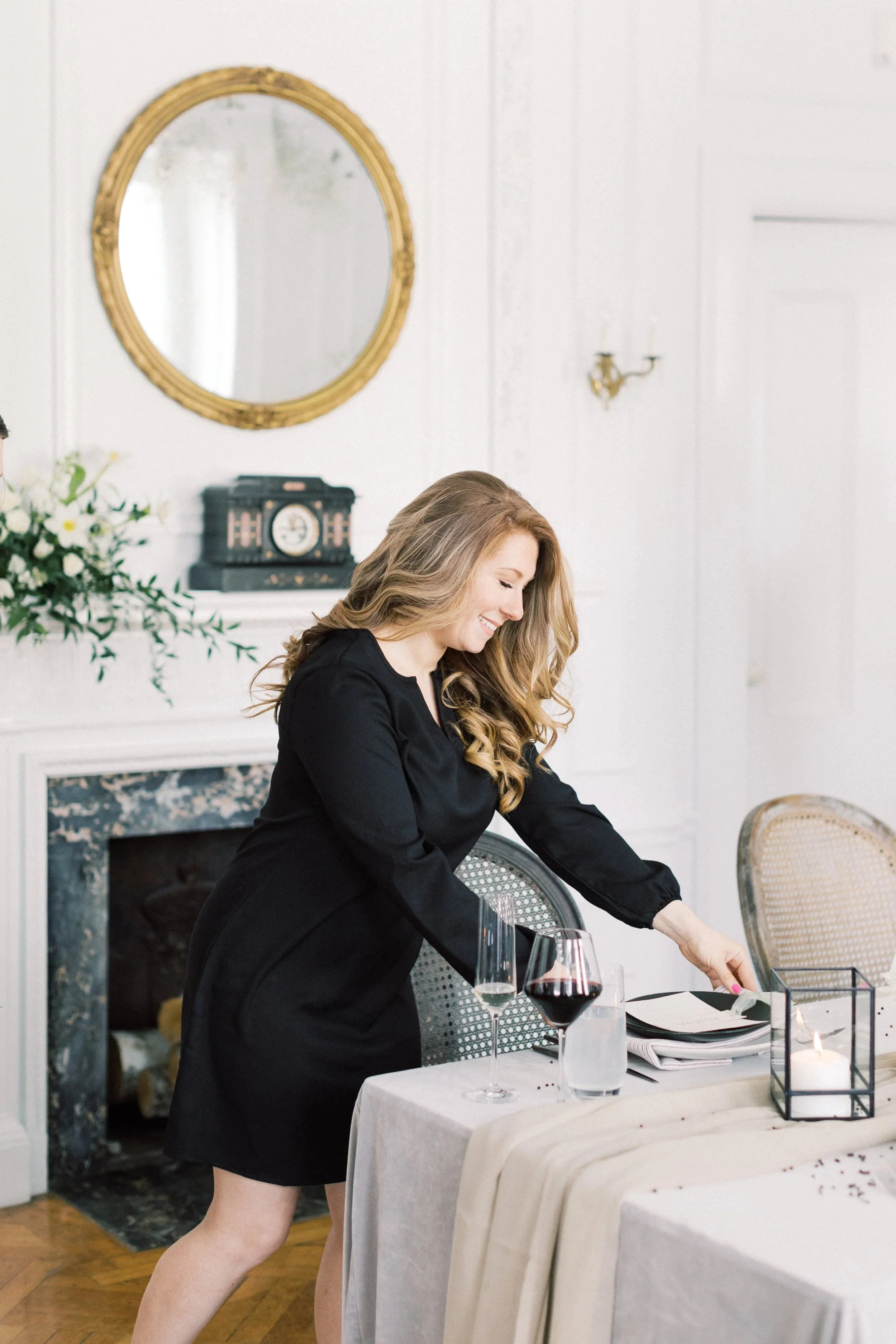 A woman in a black dress sets a table with wine glasses, a black candle holder with a white candle, and place settings in a decorated dining room.