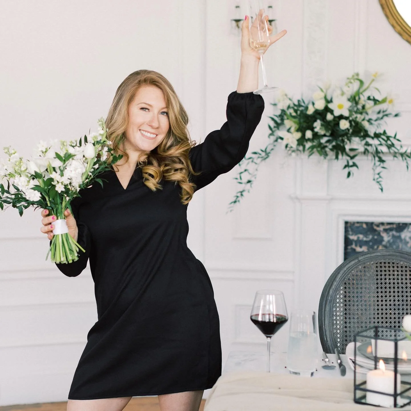 A woman with long, wavy blonde hair, wearing a black dress, smiling and holding a bouquet of white flowers in one hand and a glass of champagne raised in the other, in a decorated room with flowers and candles.