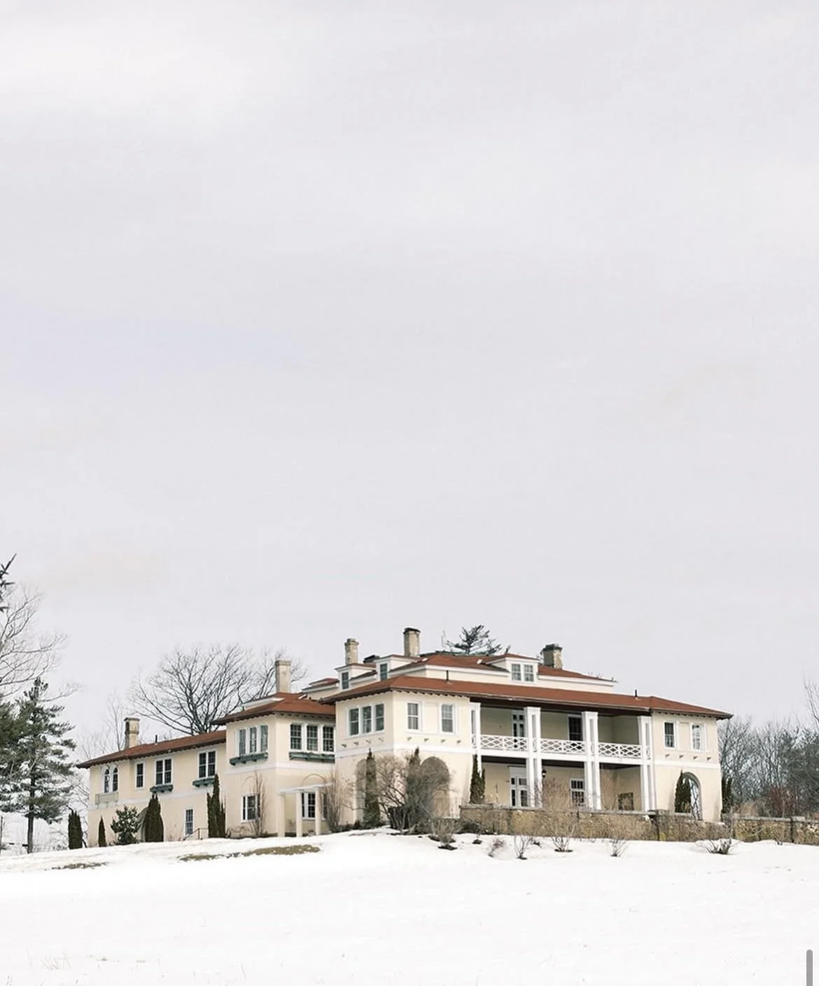 Large white mansion with multiple chimneys, surrounded by snow and trees, under an overcast sky.