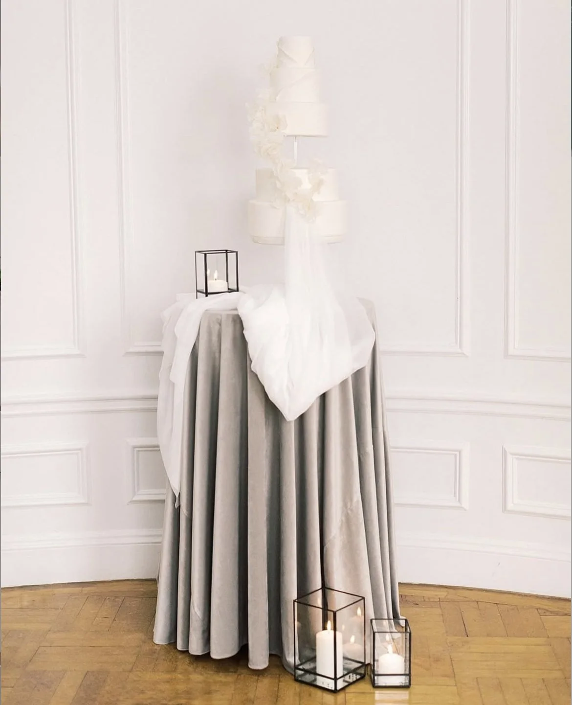 Elegant table with a gray velvet tablecloth, decorated with white fabric and candles in black metal and glass lanterns, against a white wall with decorative paneling.
