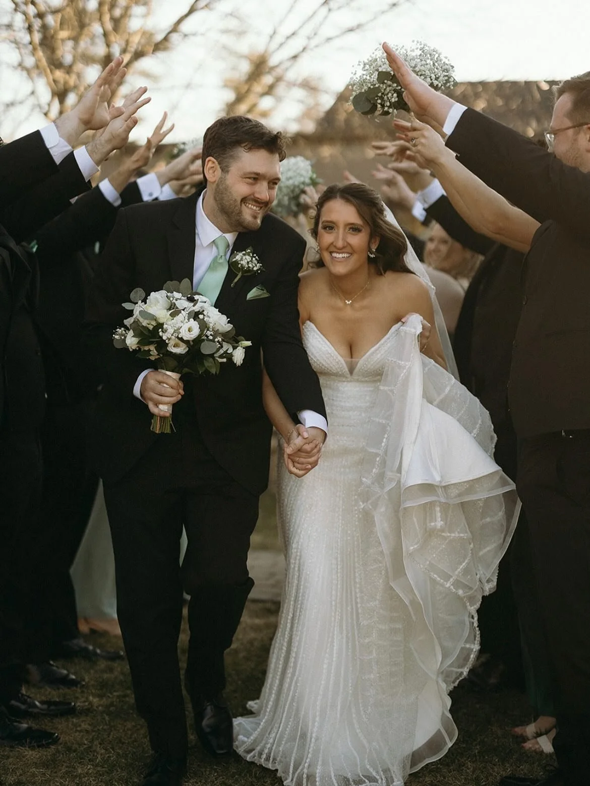 Wedding couple walking hand in hand surrounded by friends or family forming an arch. The bride is wearing a white wedding gown, and the groom is in a black suit with a light green tie. The groom is holding a bouquet and smiling, as is the bride. They are outdoors with trees and a clear sky in the background.