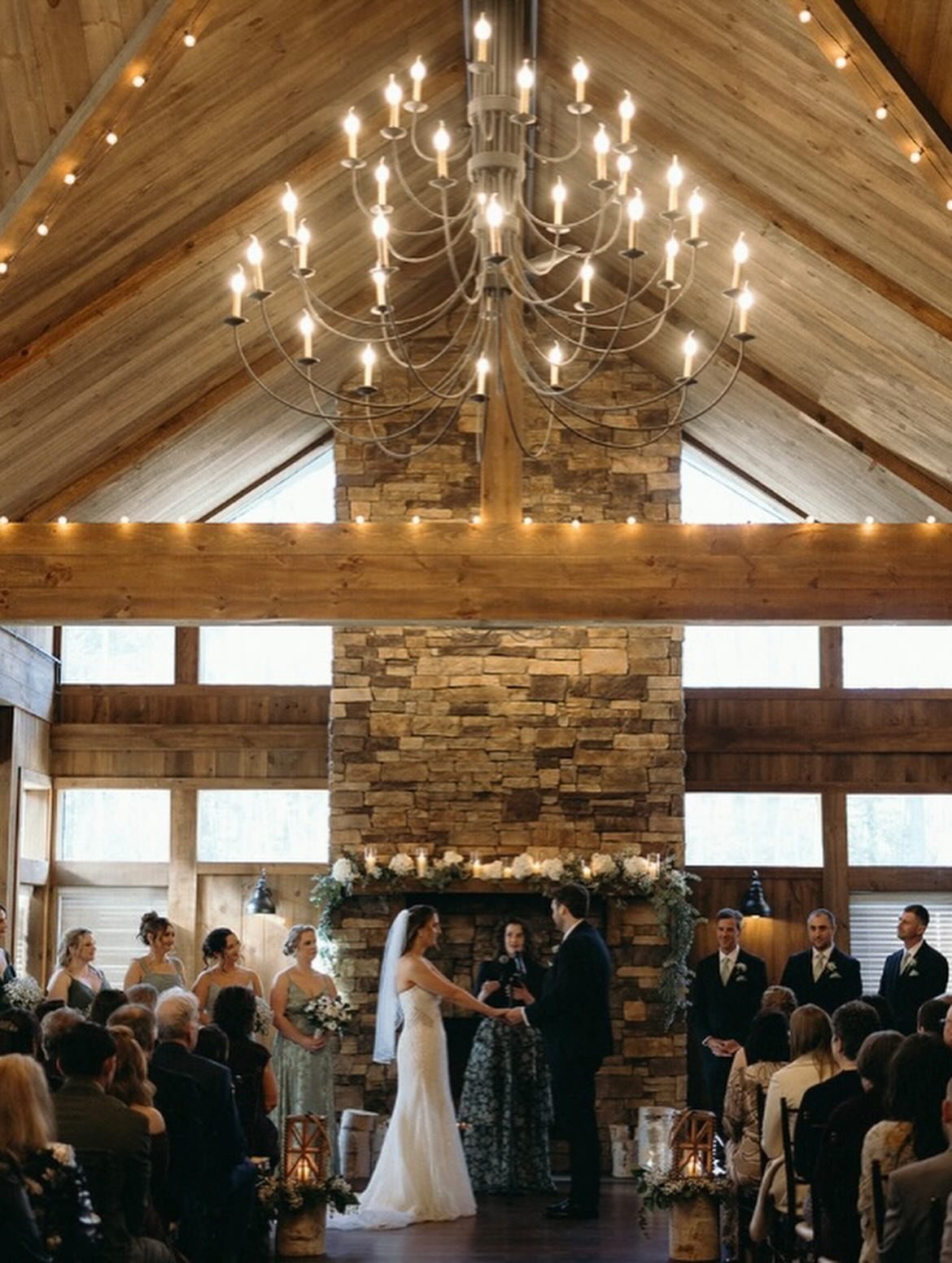 A wedding ceremony taking place in a rustic indoor venue with wood-paneled walls and a stone fireplace, a large chandelier hanging from the vaulted ceiling, and floral decorations on the fireplace. The bride and groom are standing facing each other, holding hands, with guests seated around them.