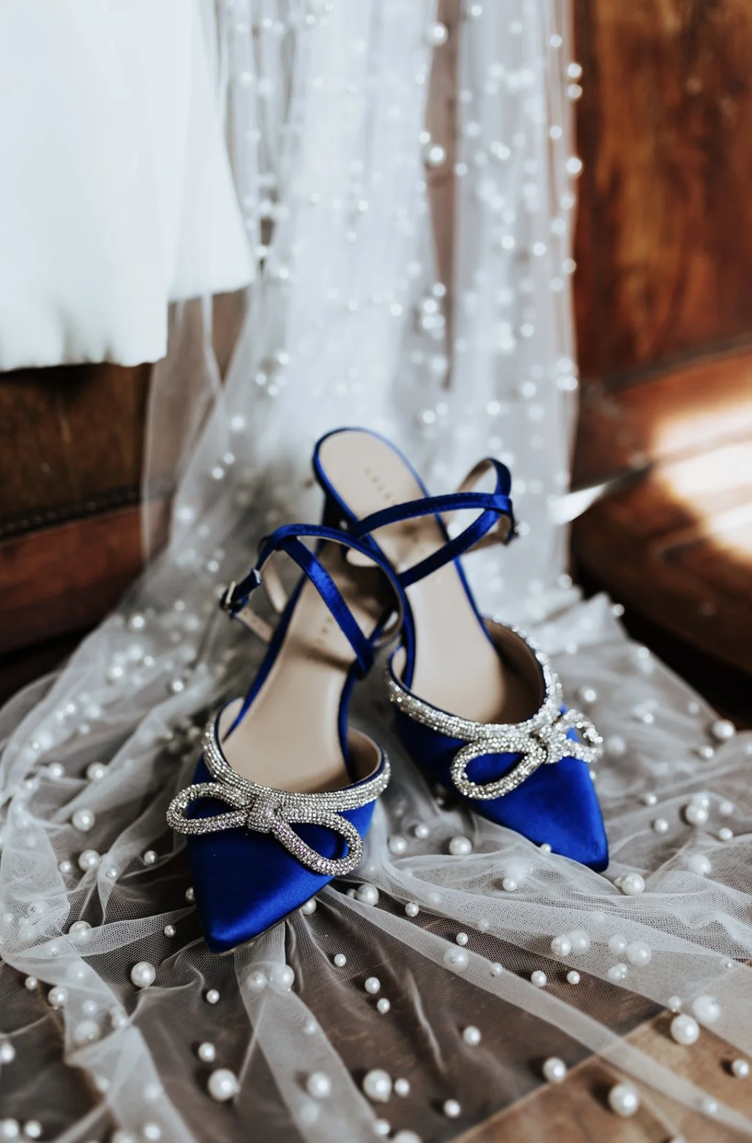 A pair of royal blue high-heeled shoes with rhinestone bows, placed on a sheer fabric decorated with pearls, next to a wedding dress.