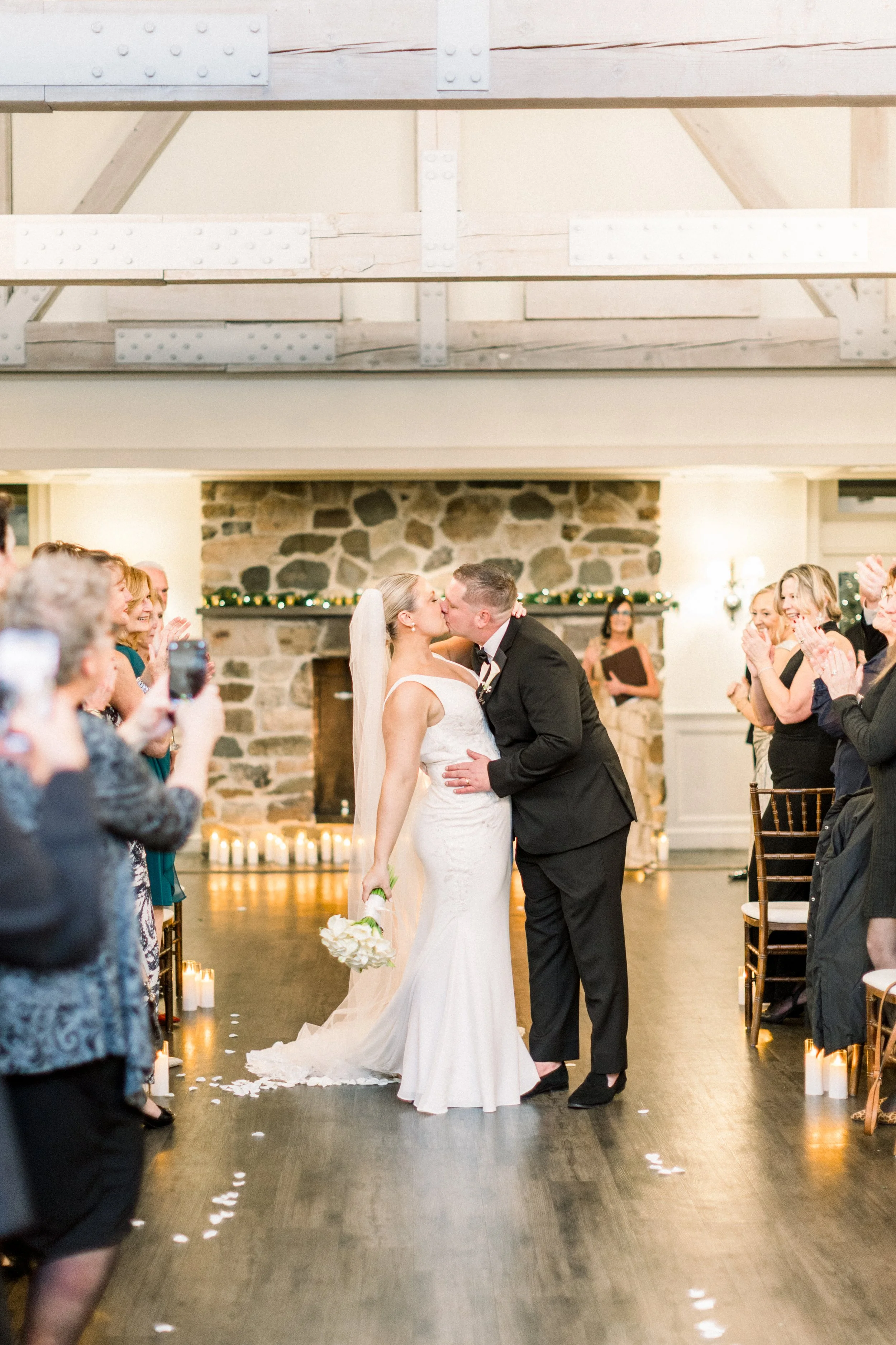 Bride and groom sharing a kiss at their wedding ceremony with guests applauding around them in a decorated indoor venue.