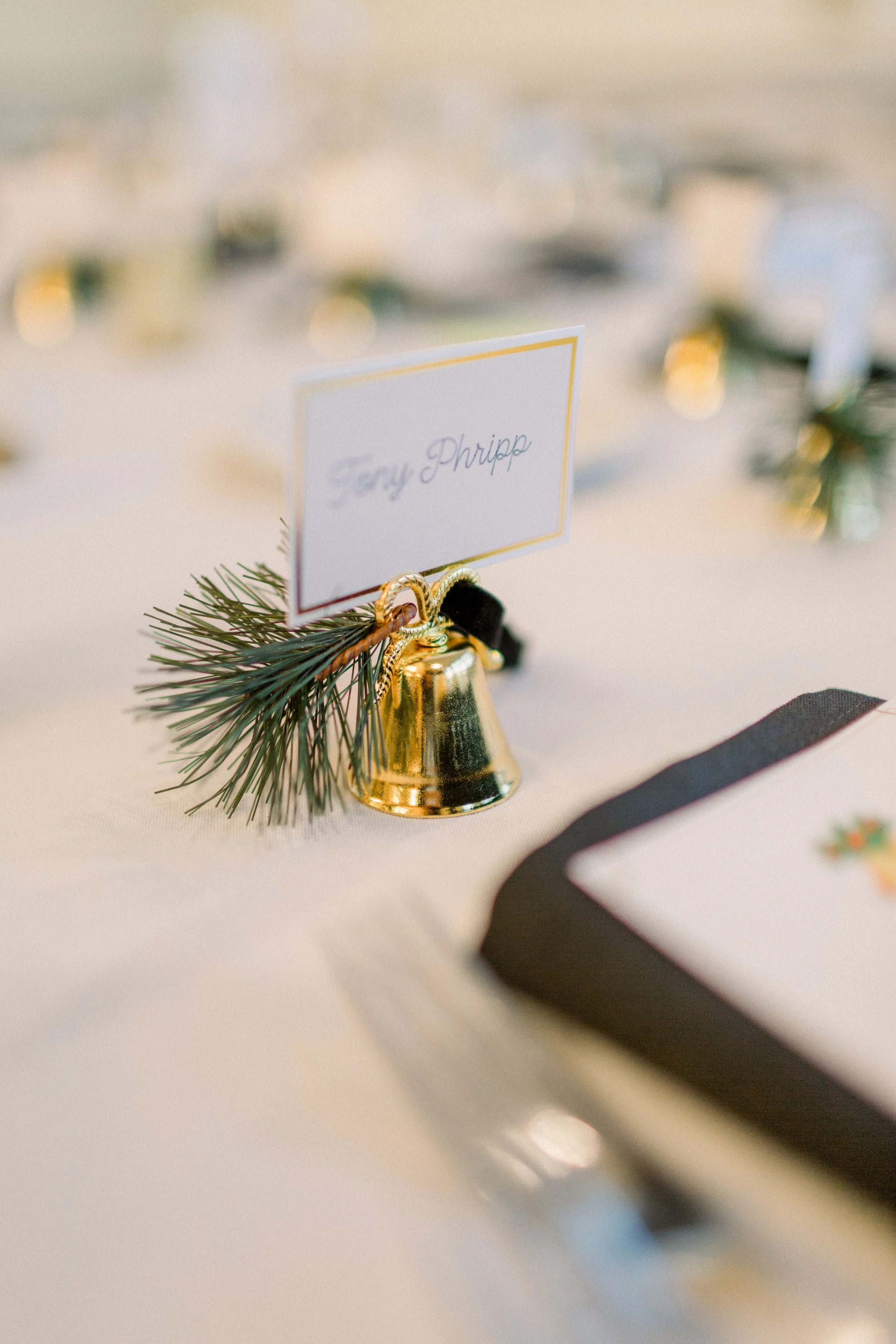 A gold decorative bell with a small pine branch and a white place card on a table with holiday decorations for a wedding celebration.