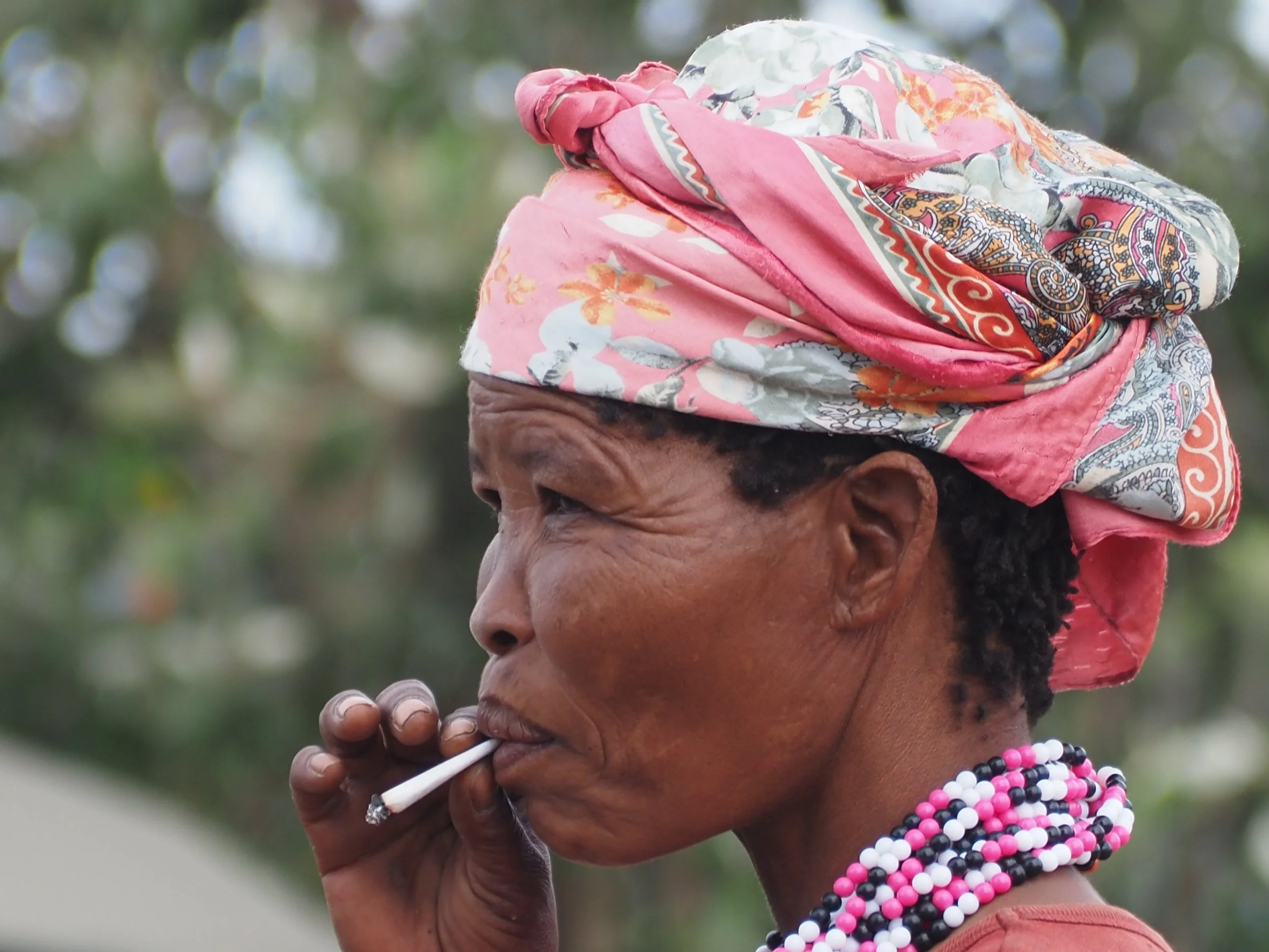 A woman with dark skin and possibly indigenous heritage, dressed in traditional clothing and wearing layered beaded necklaces, is seen smoking a cigarette outdoors with a blurred green background.