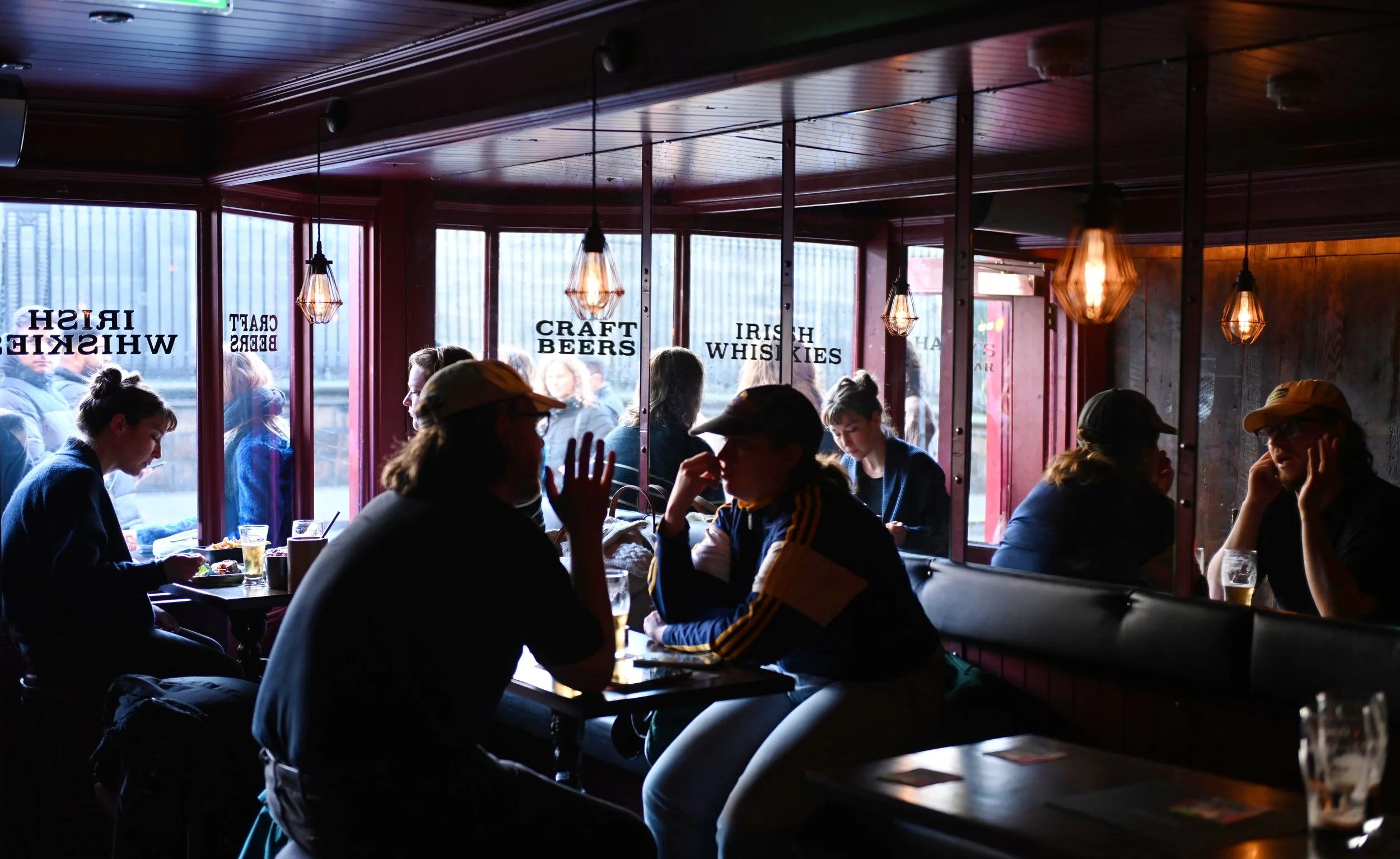 People sitting inside a pub or bar, some engaged in conversations, with large windows showing a cityscape outside. Decor includes hanging warm lights and words on the glass such as 'Irish Whiskies,' 'Craft Beers,' and 'Irish Whiskey'.