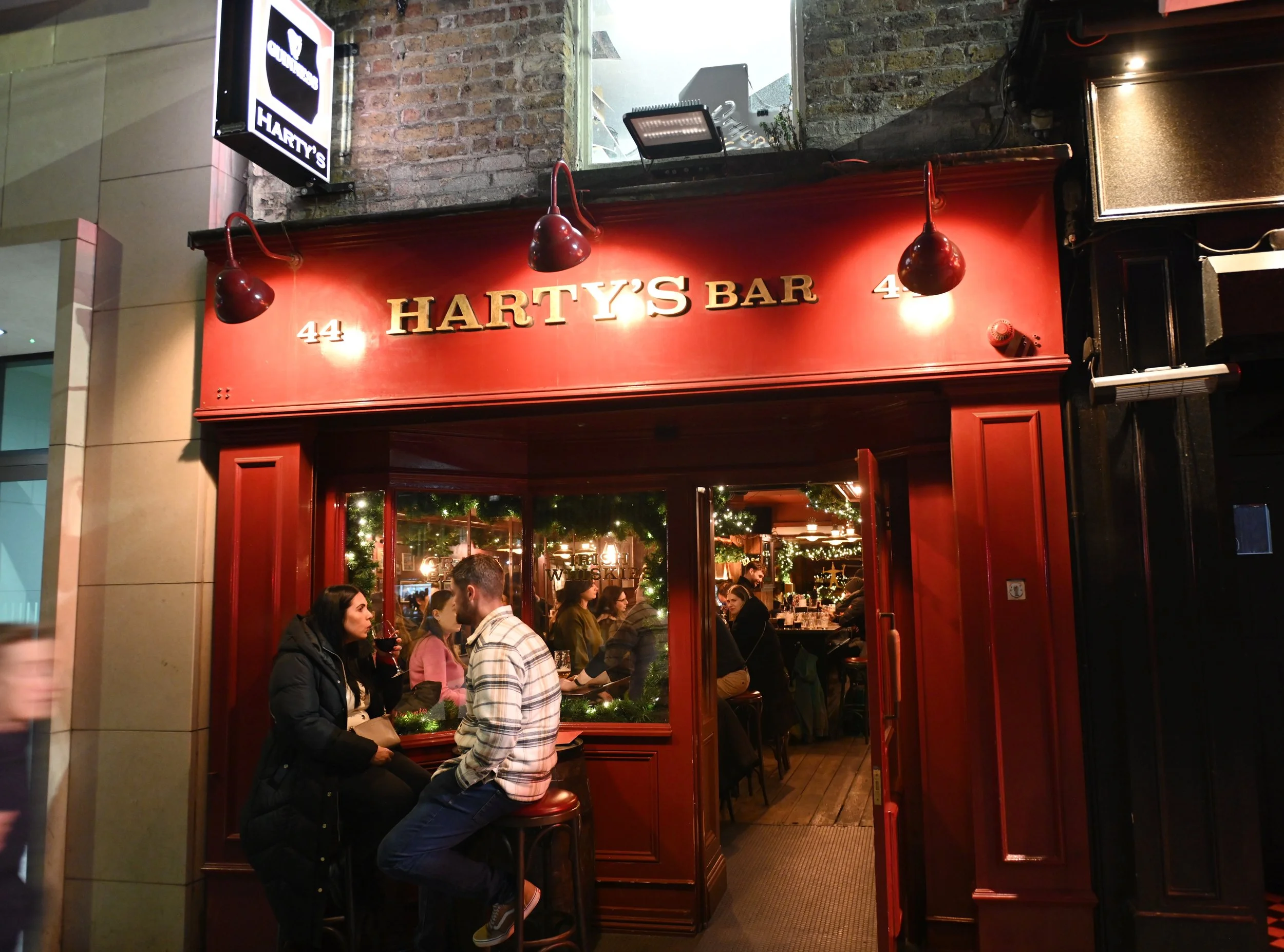 Exterior view of Harty's Bar at night with a red facade, showing the bar's name and open door revealing people inside, with some customers sitting outside at a small table.