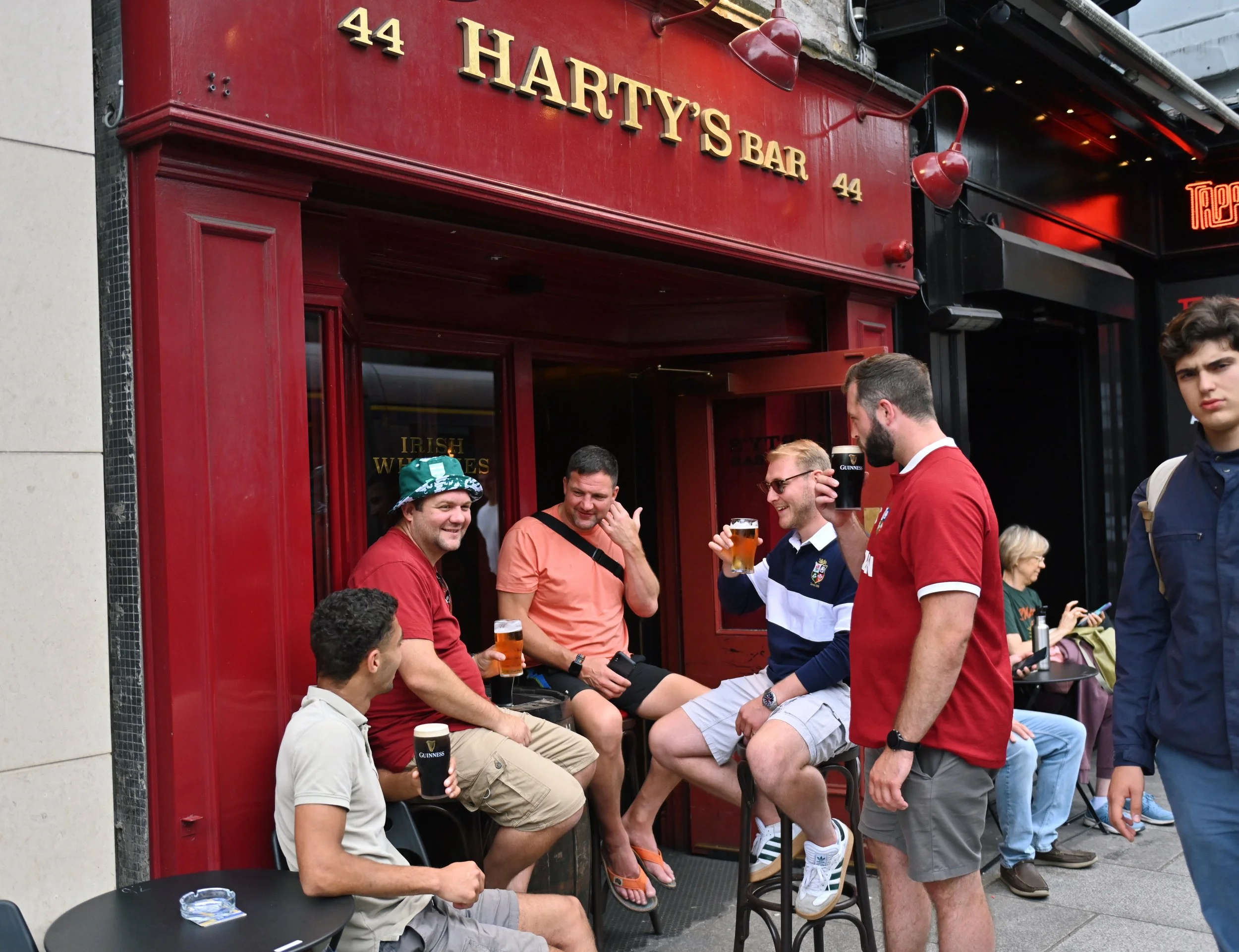 A group of men with drinks enjoying outdoor seating outside Harty's Bar, with a person in the background on the sidewalk.