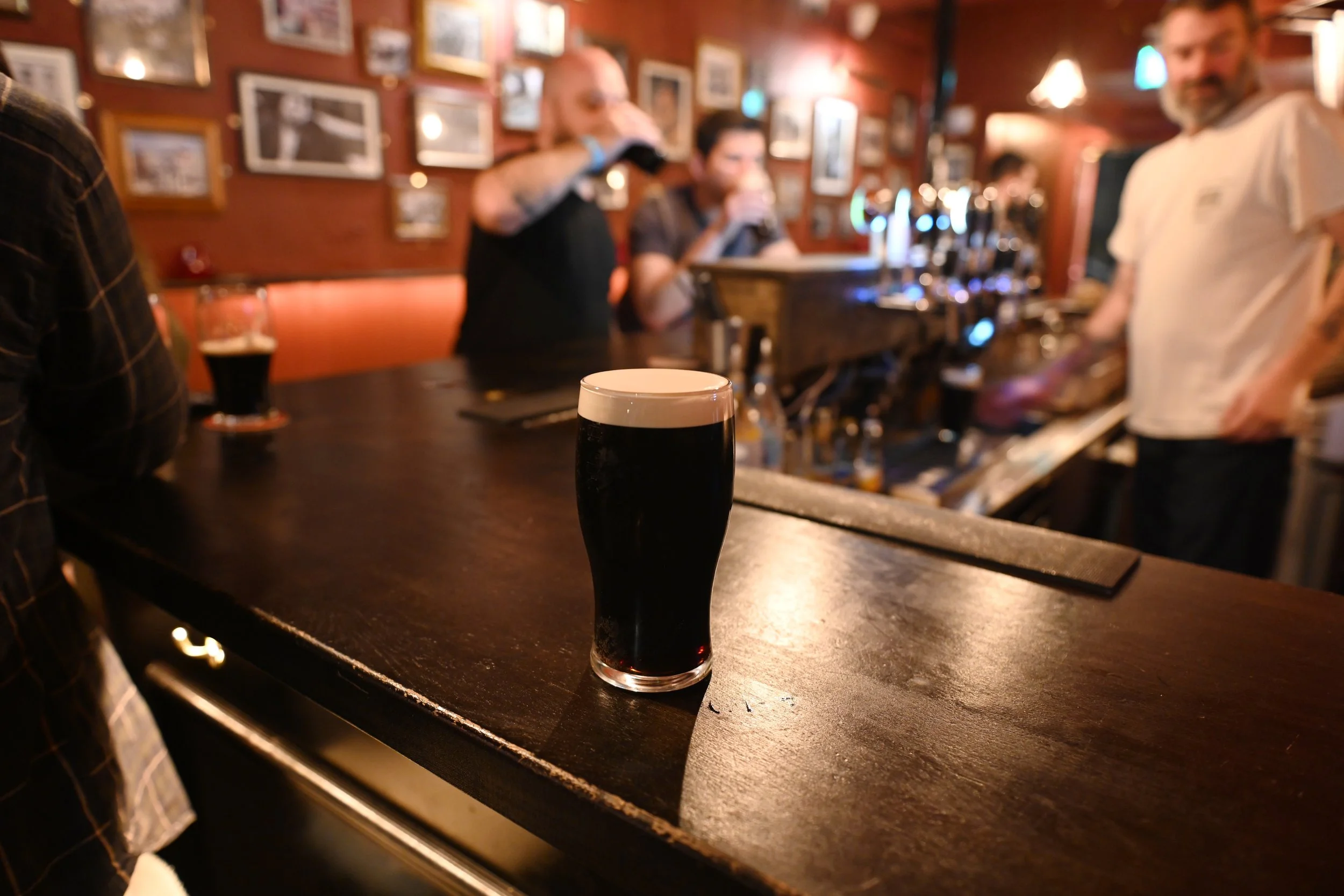 A pint glass of stout with a creamy head on a wooden bar counter in a dimly lit pub, with beer taps and bartenders in the background.