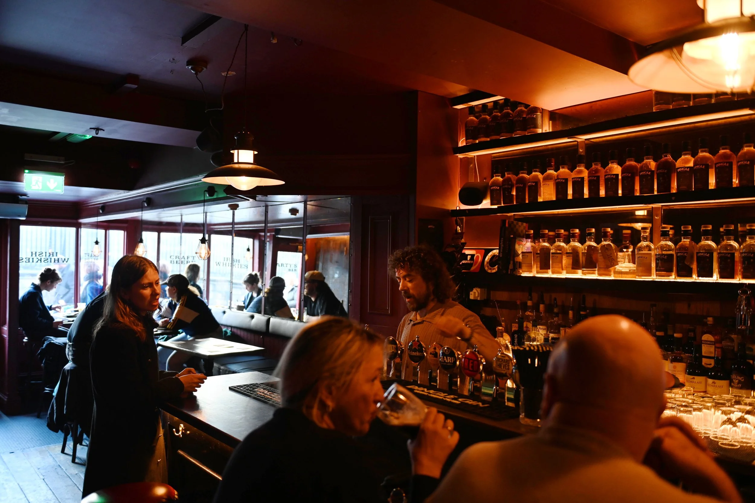 People sitting at a bar in a dimly lit pub, with shelves of liquor bottles behind the bartender and patrons drinking and socializing.