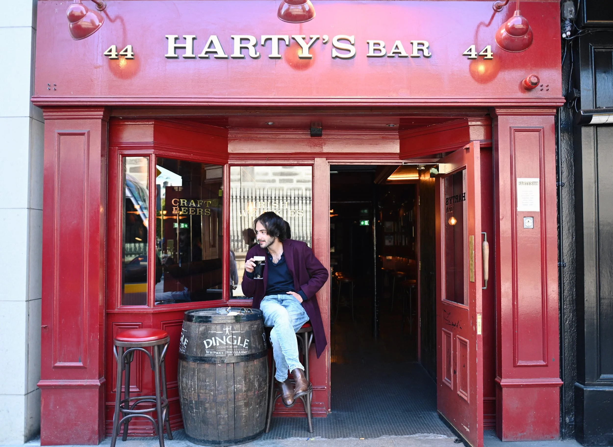 A man sitting outside a red bar named Harty's Bar, drinking a dark beverage. The bar entrance has a large sign with the name and the number 44. The man is seated on a stool next to a barrel, with a stool beside him. The interior of the bar is dimly l